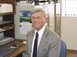 Man in a suit smiles while sitting at a desk with a computer, drawers, and a calendar.