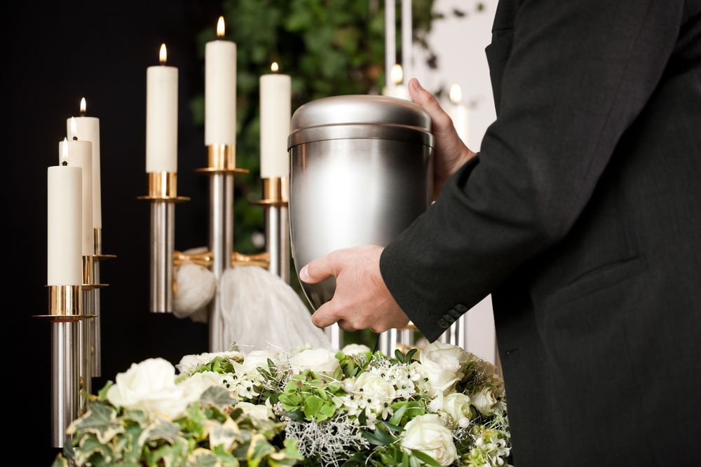 Person in black suit holding an urn at a memorial. Candles and white flowers surround.