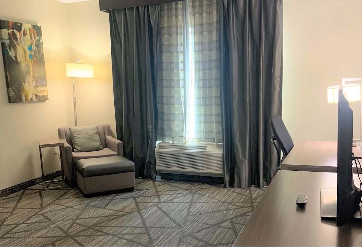 A hotel room with a window, armchair, desk and artwork. Dark gray curtains and a patterned carpet.