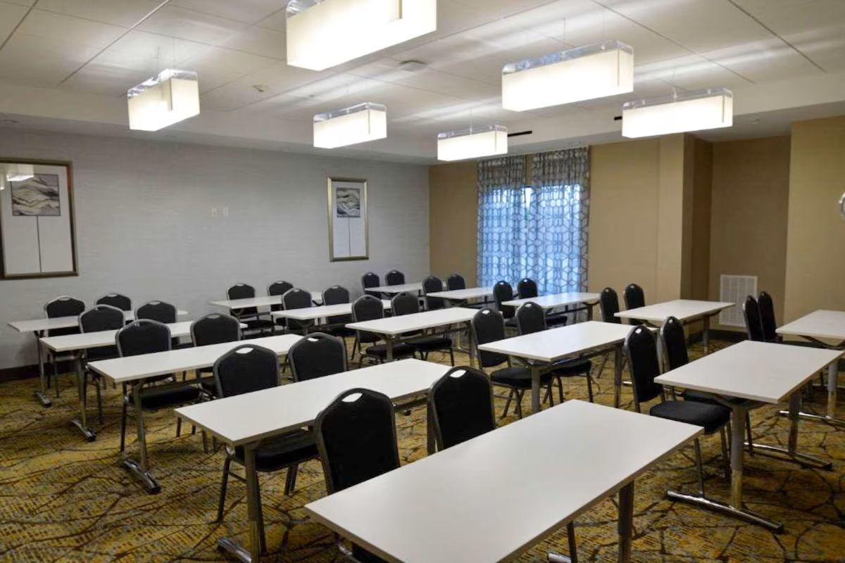 Classroom with tables and chairs arranged in rows; beige walls, patterned carpet, rectangular lights.
