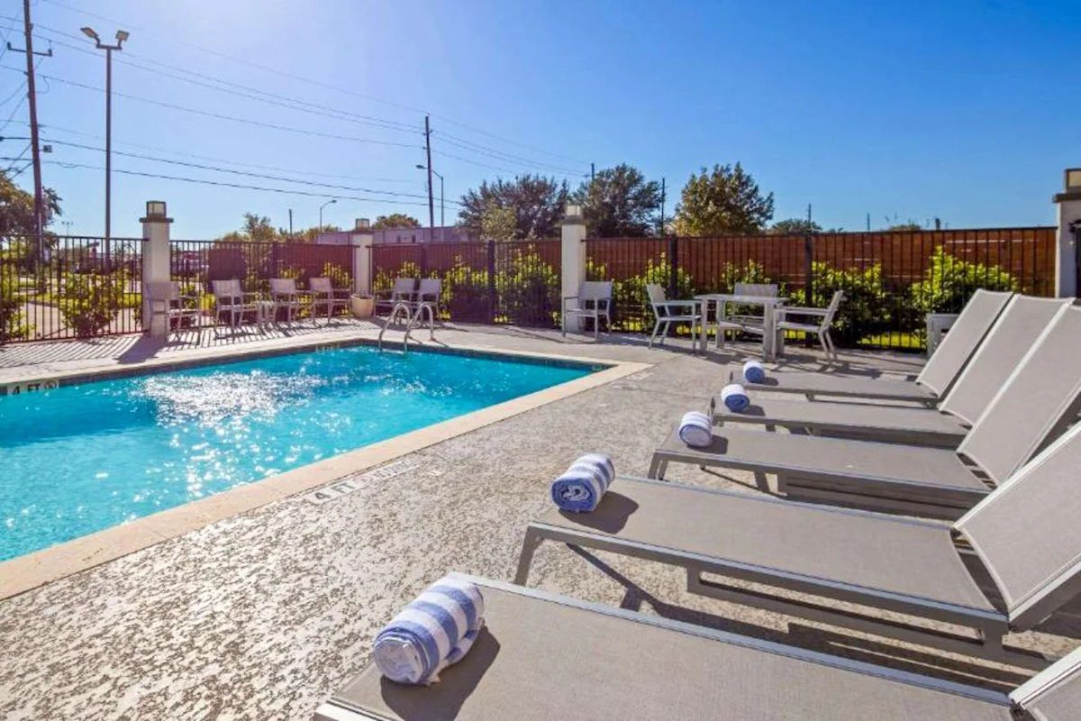 Poolside scene with lounge chairs, blue water, and tables under a sunny sky.