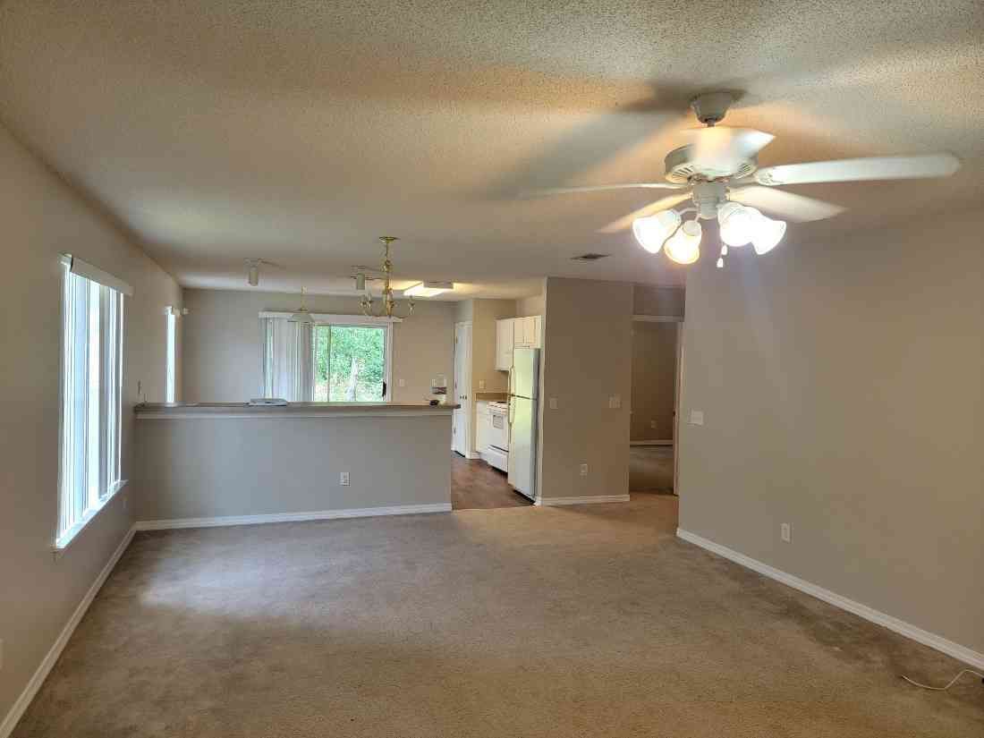 An empty living room with a ceiling fan and a kitchen in the background.
