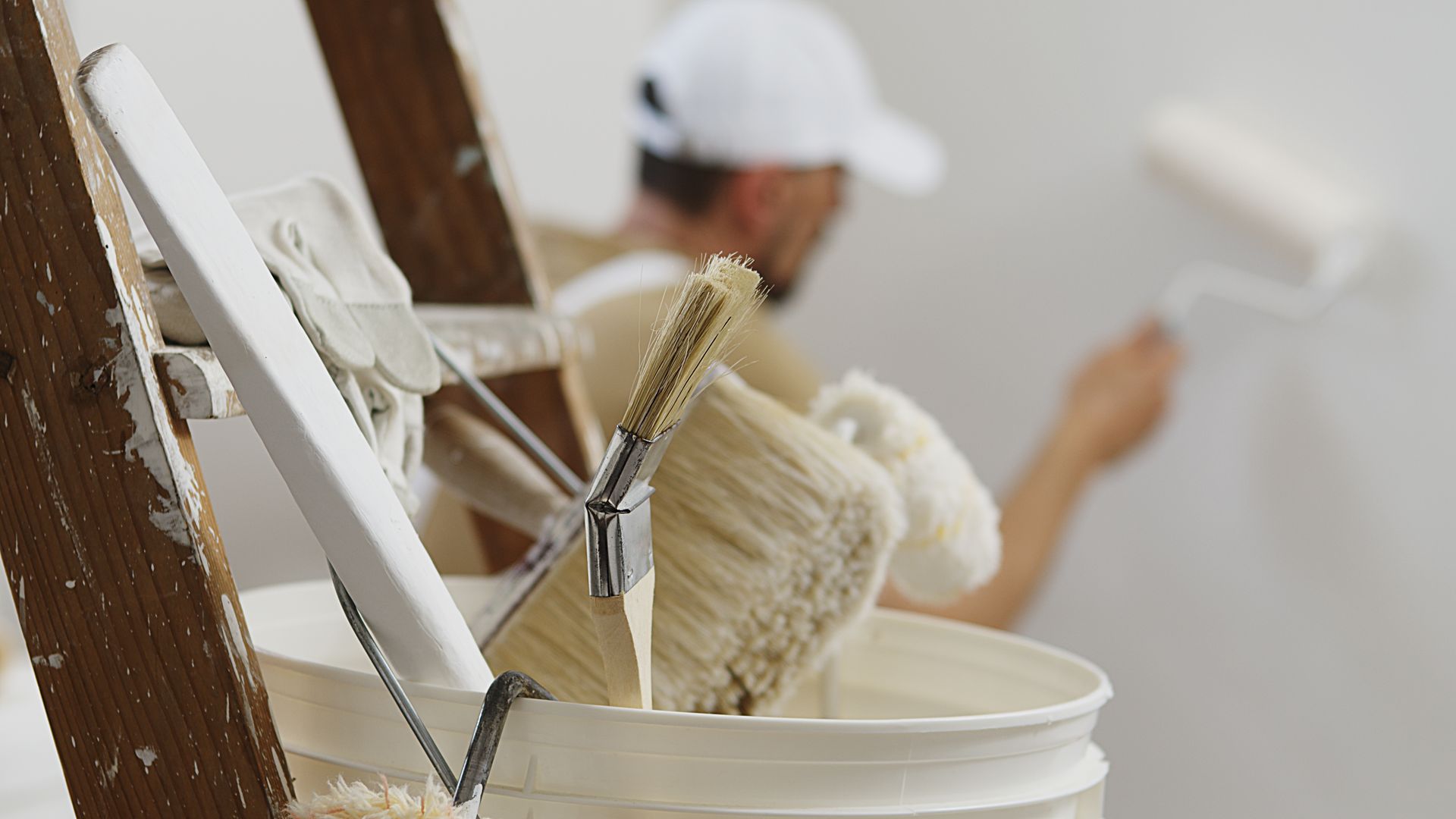 Painter in white cap rolling paint on a wall, brushes and paint bucket in the foreground.