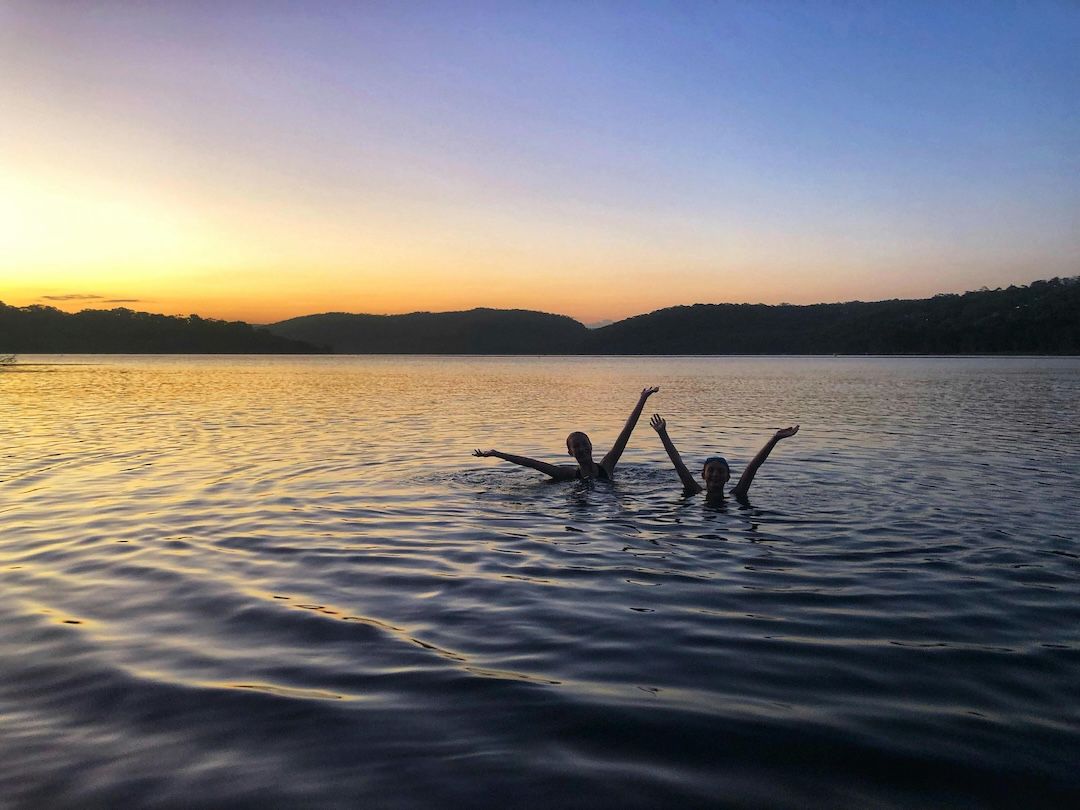 Two silhouetted people swimming and raising their arms in a calm lake reflecting a bright orange sunset.