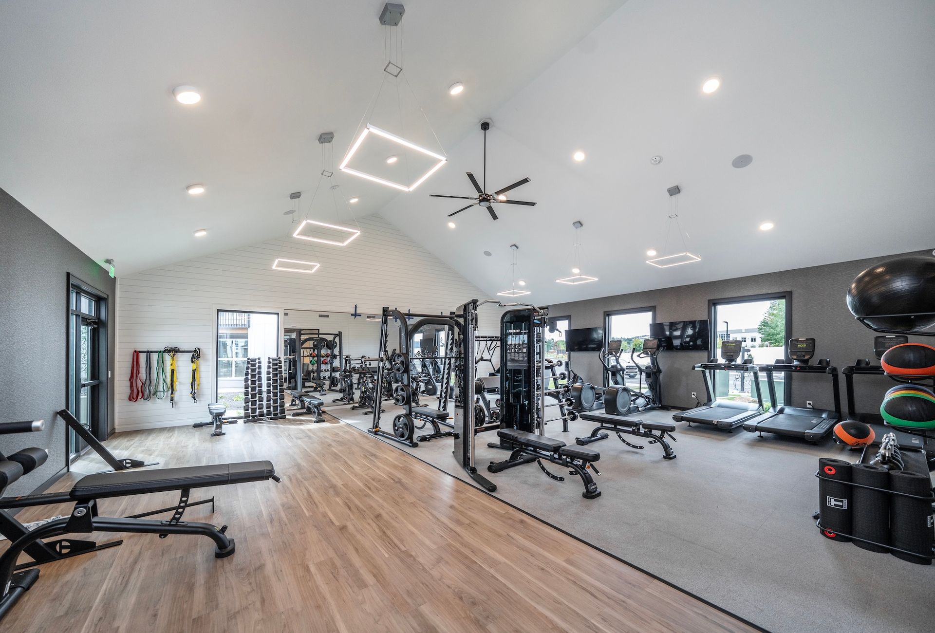 Bright, spacious gym with weight machines, benches, and treadmills. Wooden floor, gray walls, and modern lighting.