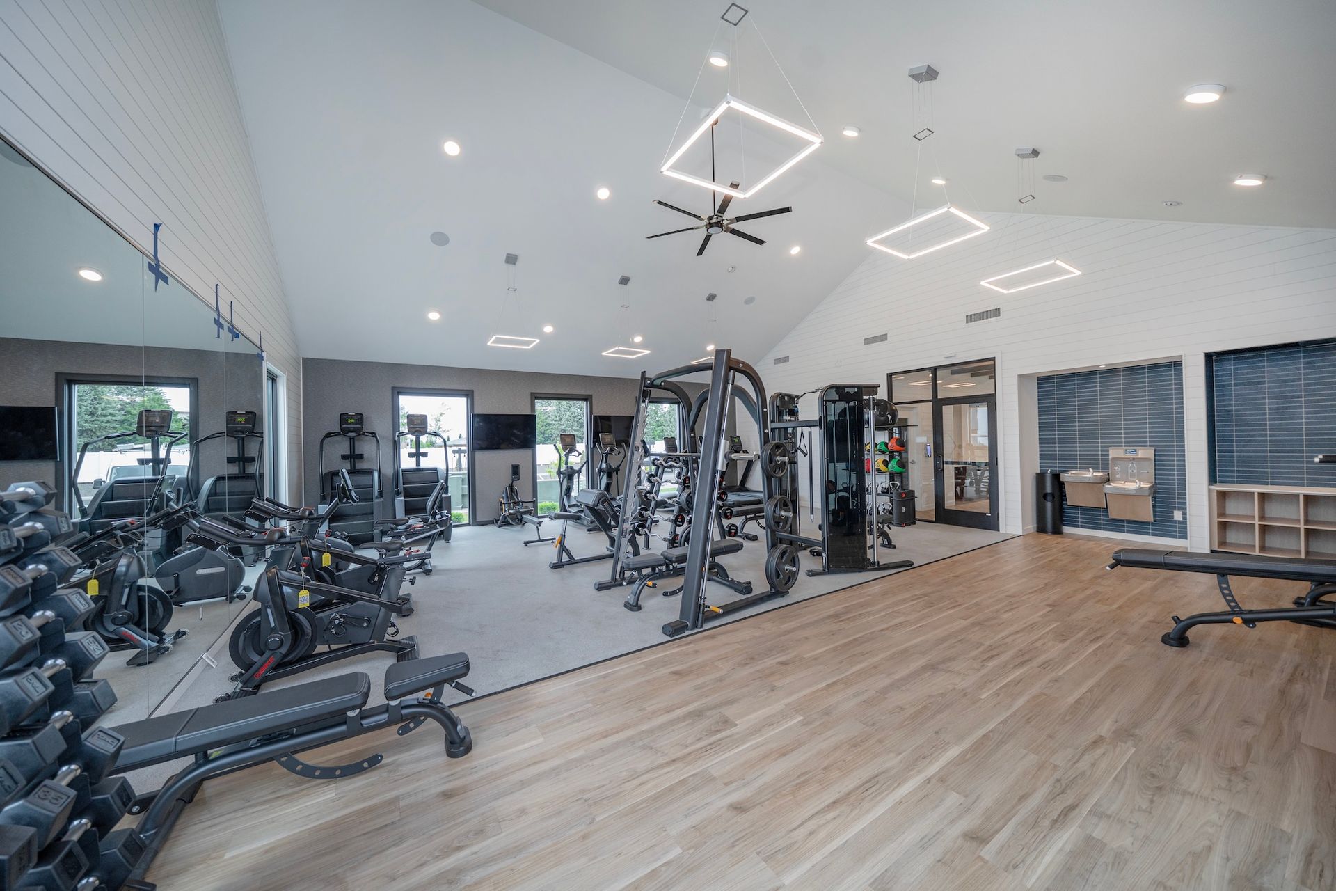 Bright, modern gym with weight machines, exercise bikes, free weights, and a bench. Light wood floors.