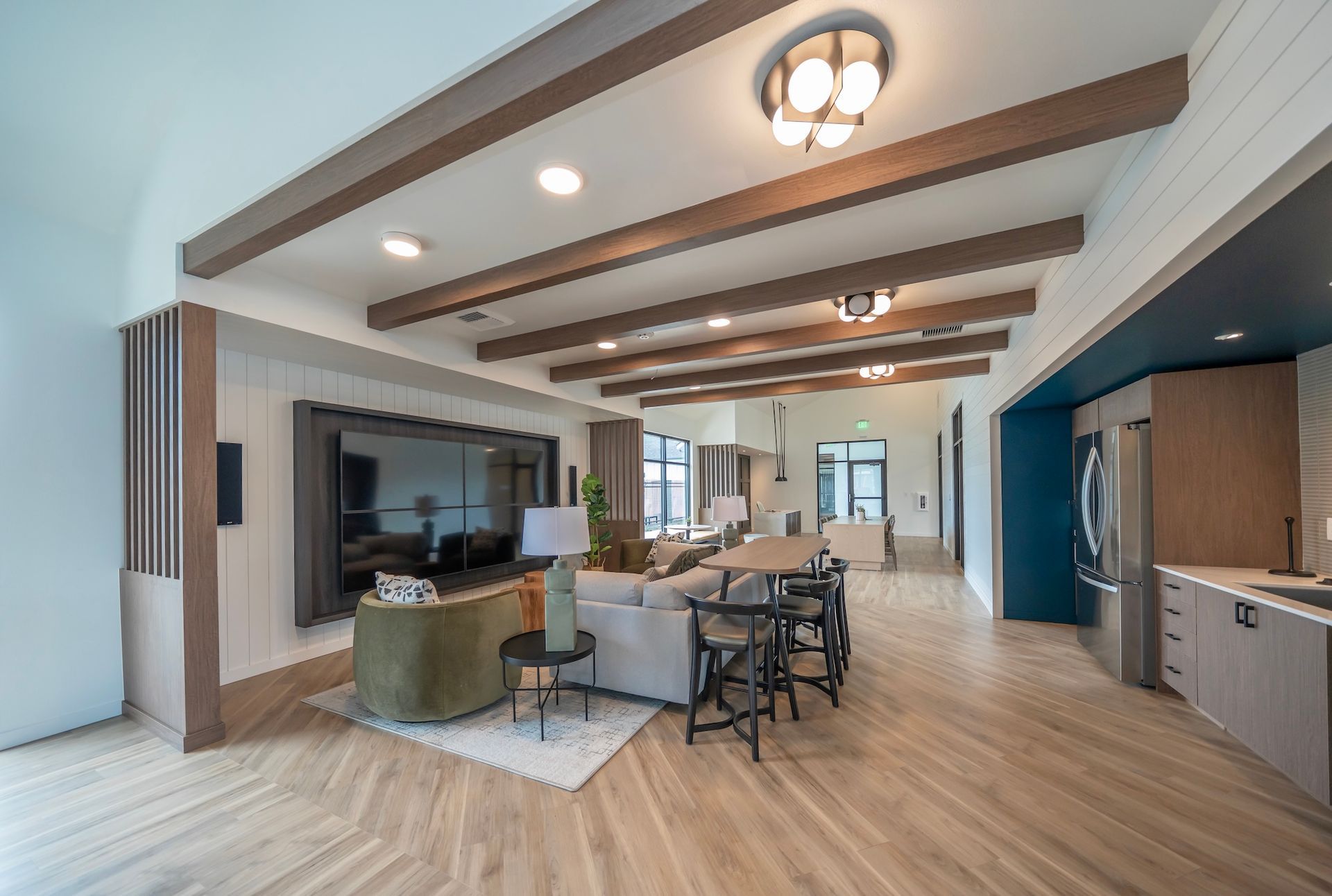 Spacious living area with wood beams, a large TV, sofa, and kitchen, lit by recessed and overhead lights.