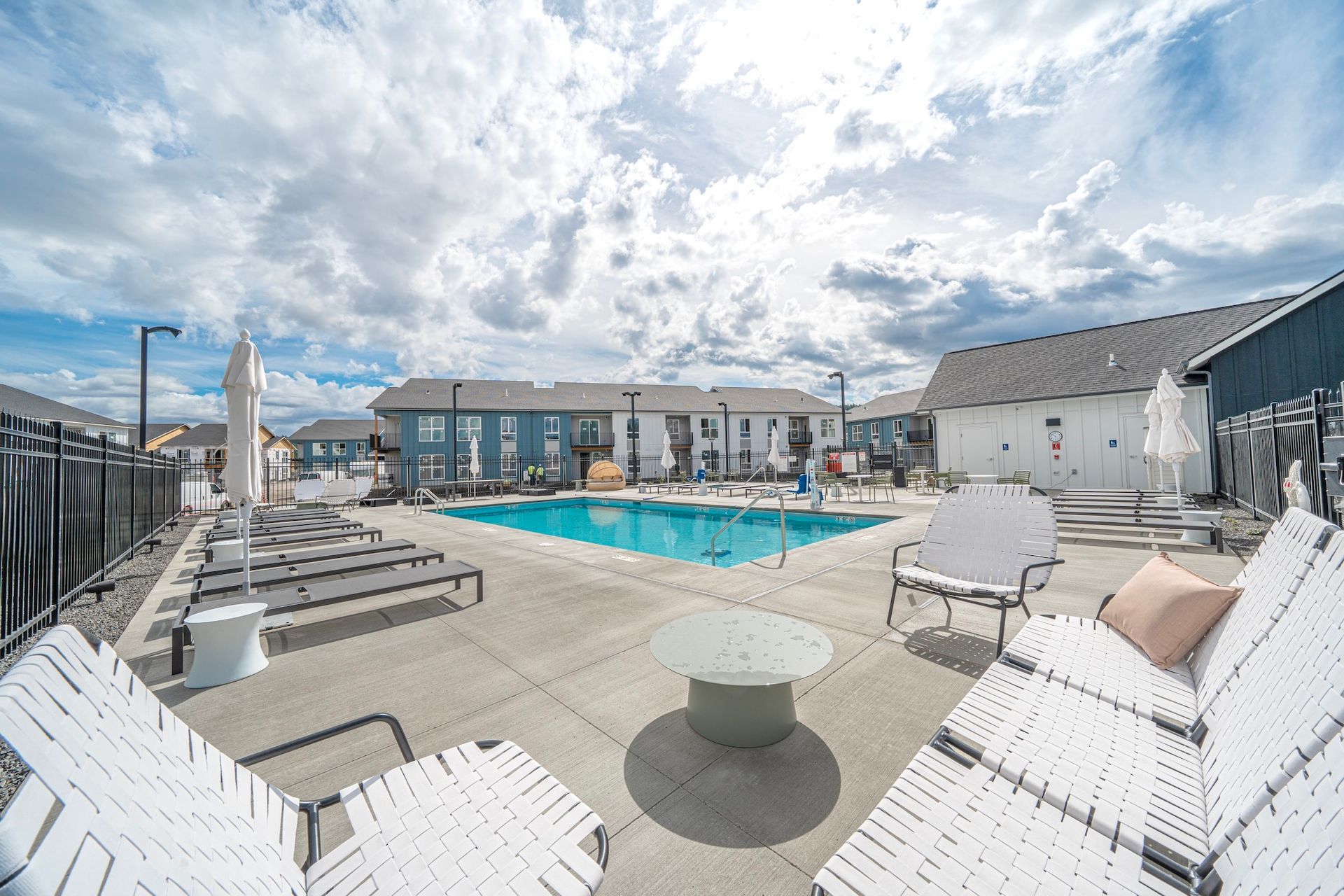 Pool area with lounge chairs, blue water, and apartment buildings under a cloudy sky.