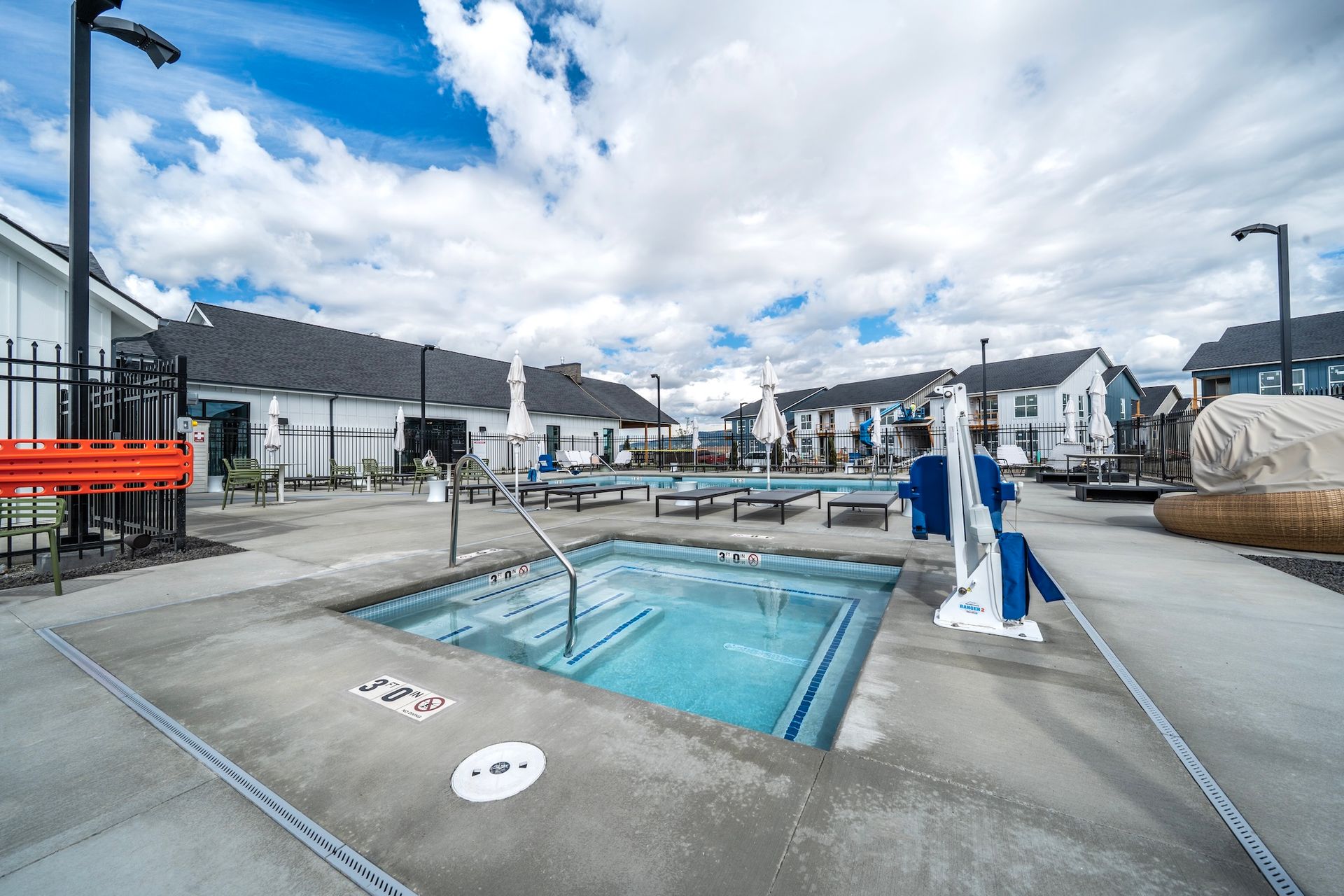 Pool area with a hot tub, blue water, and buildings in the background on a sunny day.