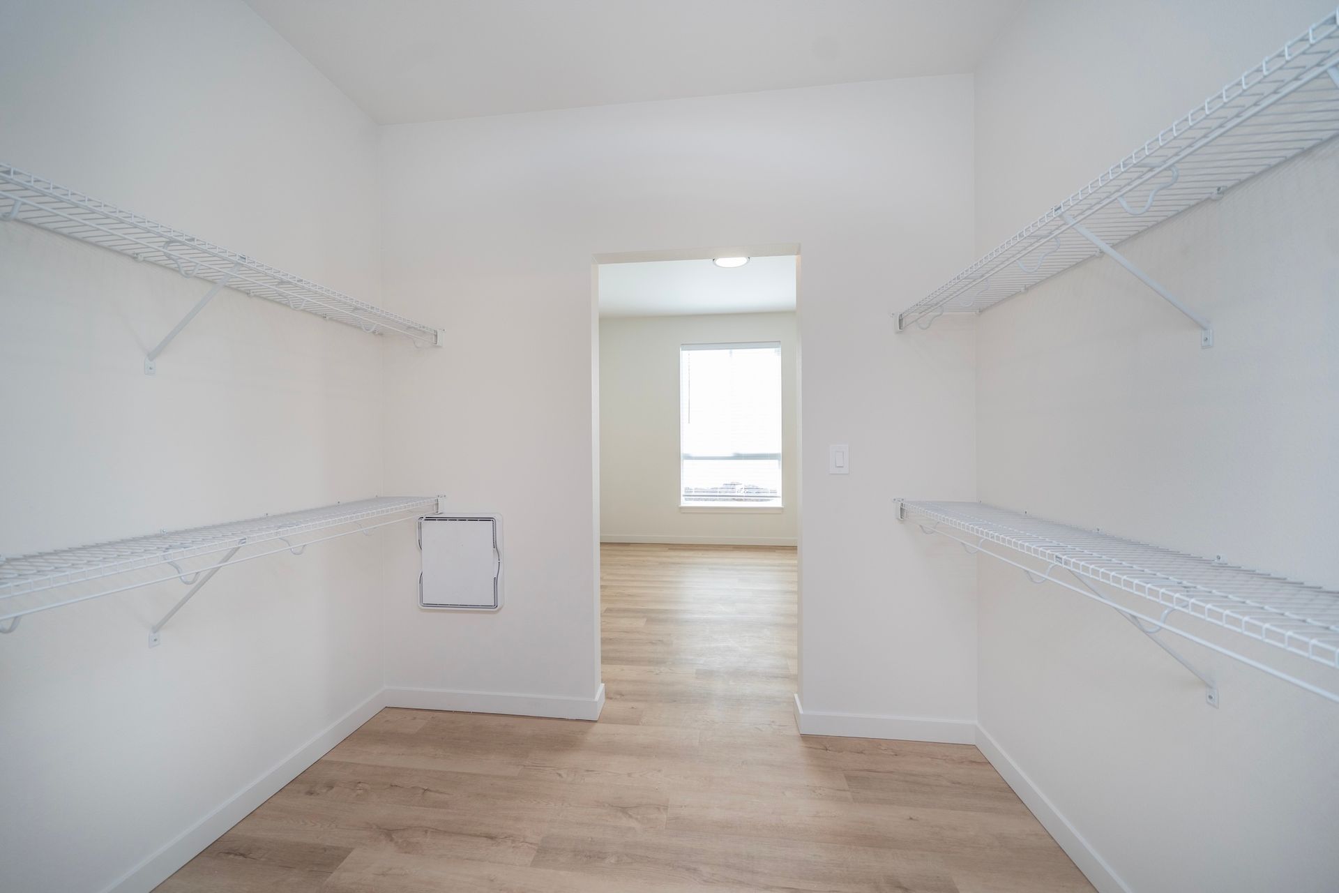 Empty walk-in closet with white wire shelves, white walls, and a doorway to a light-filled room with wood floors.