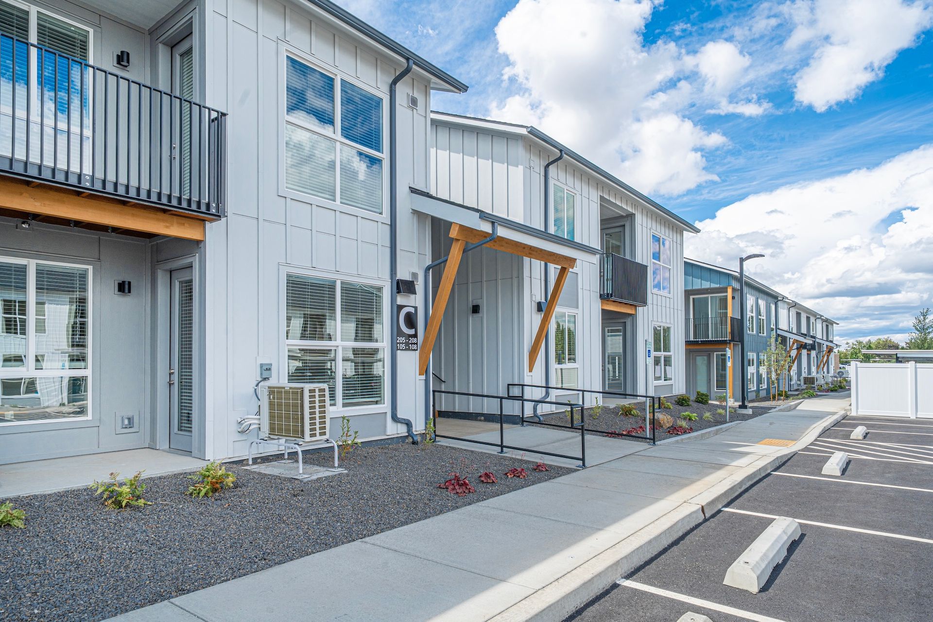 Modern gray townhomes with balconies, a walkway, and parking on a sunny day.
