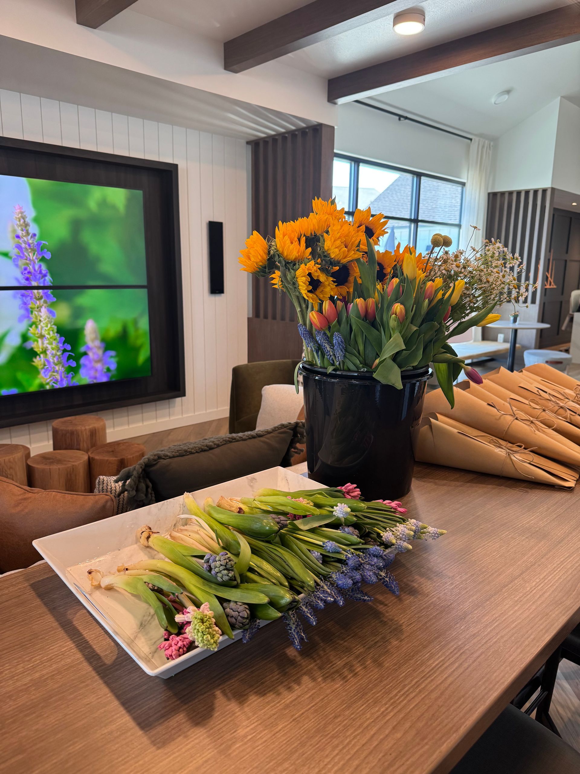 Sunflowers and tulips in a black vase and a tray on a wooden table in a brightly lit interior space.