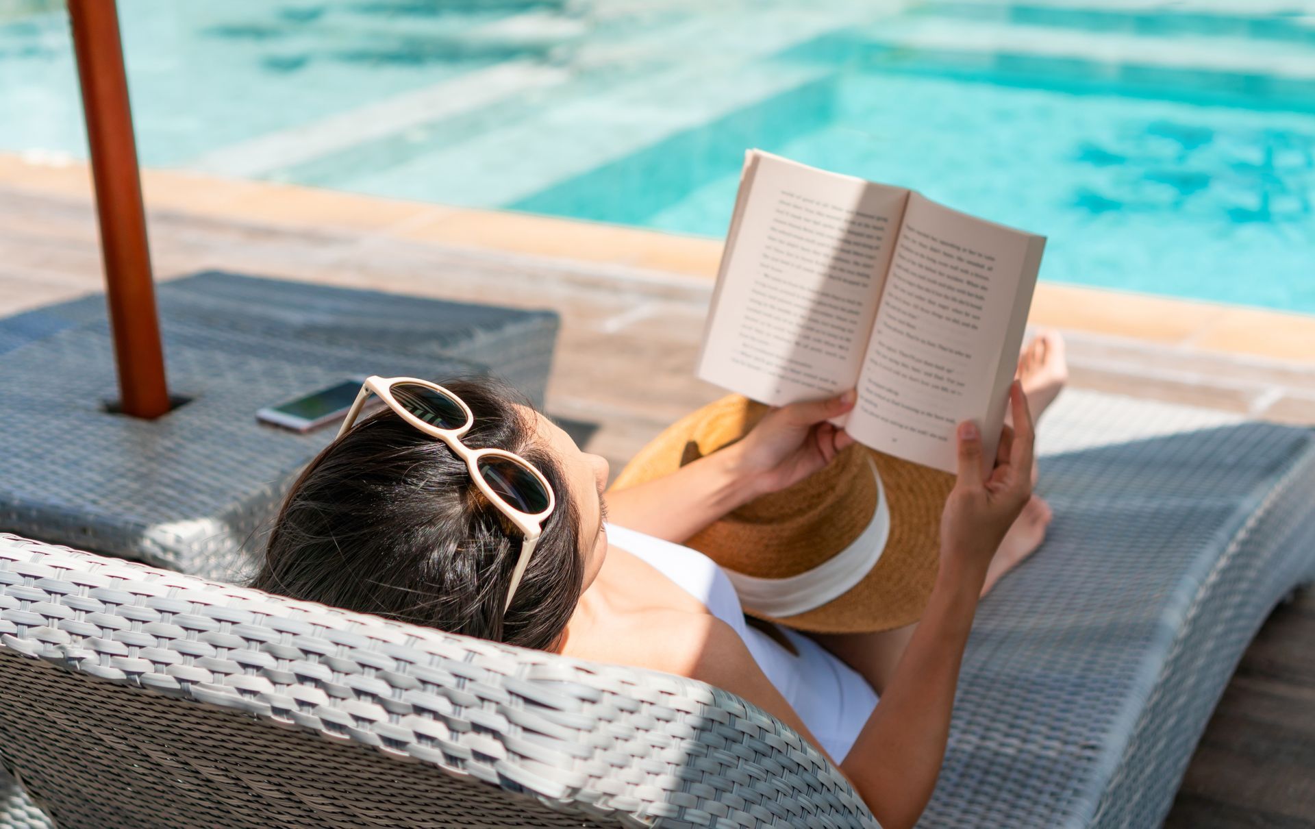 A woman is laying on a lounge chair reading a book by a pool.