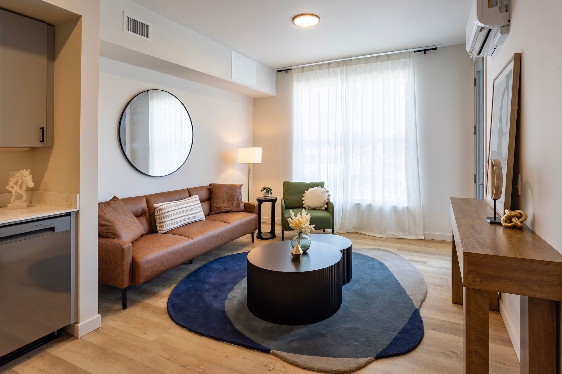Living room with a brown leather sofa, green chair, and round coffee table on a blue and gray rug. White walls and light wood floors.