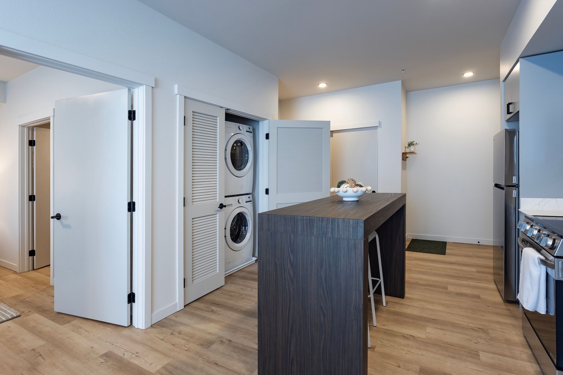 A modern kitchen area with light wood floors, a dark wood island, and stacked washer and dryer units in a closet.