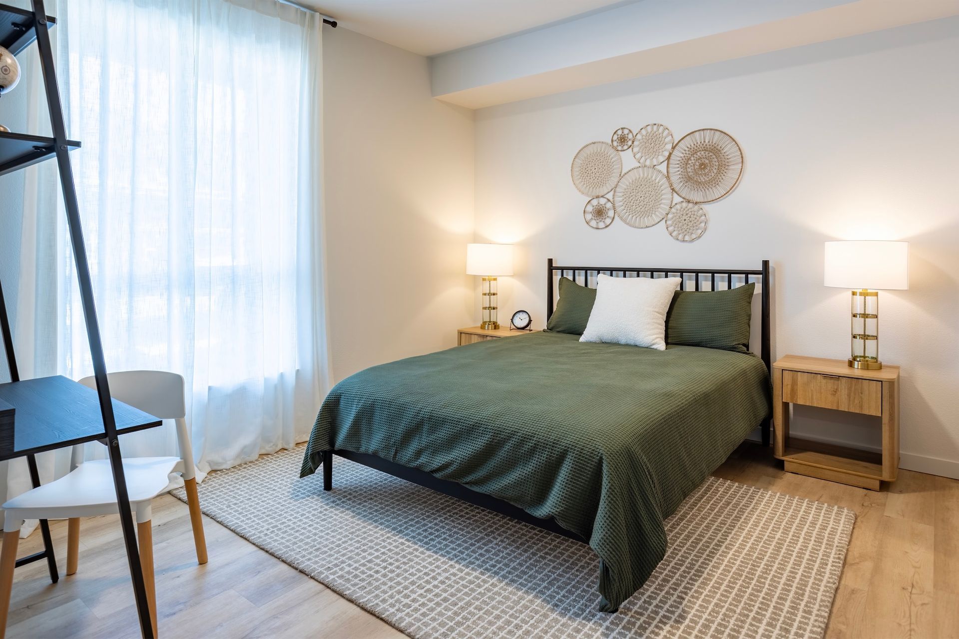 Bedroom with green bedding, two nightstands with lamps, and woven wall decor. A desk and chair are on the left.