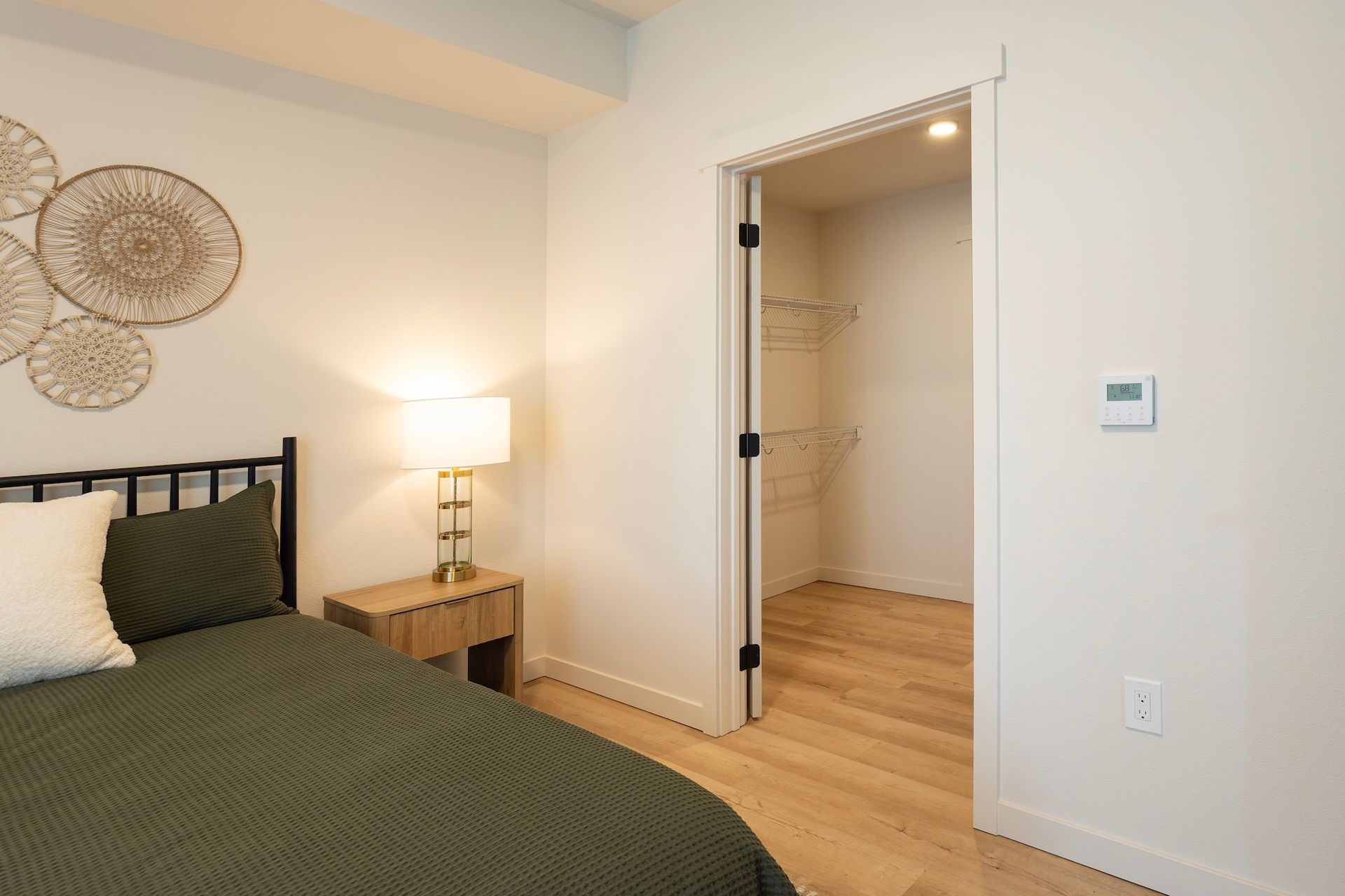A bedroom with a bed, nightstand, and closet. Olive green bedspread, light wood floors, and white walls.