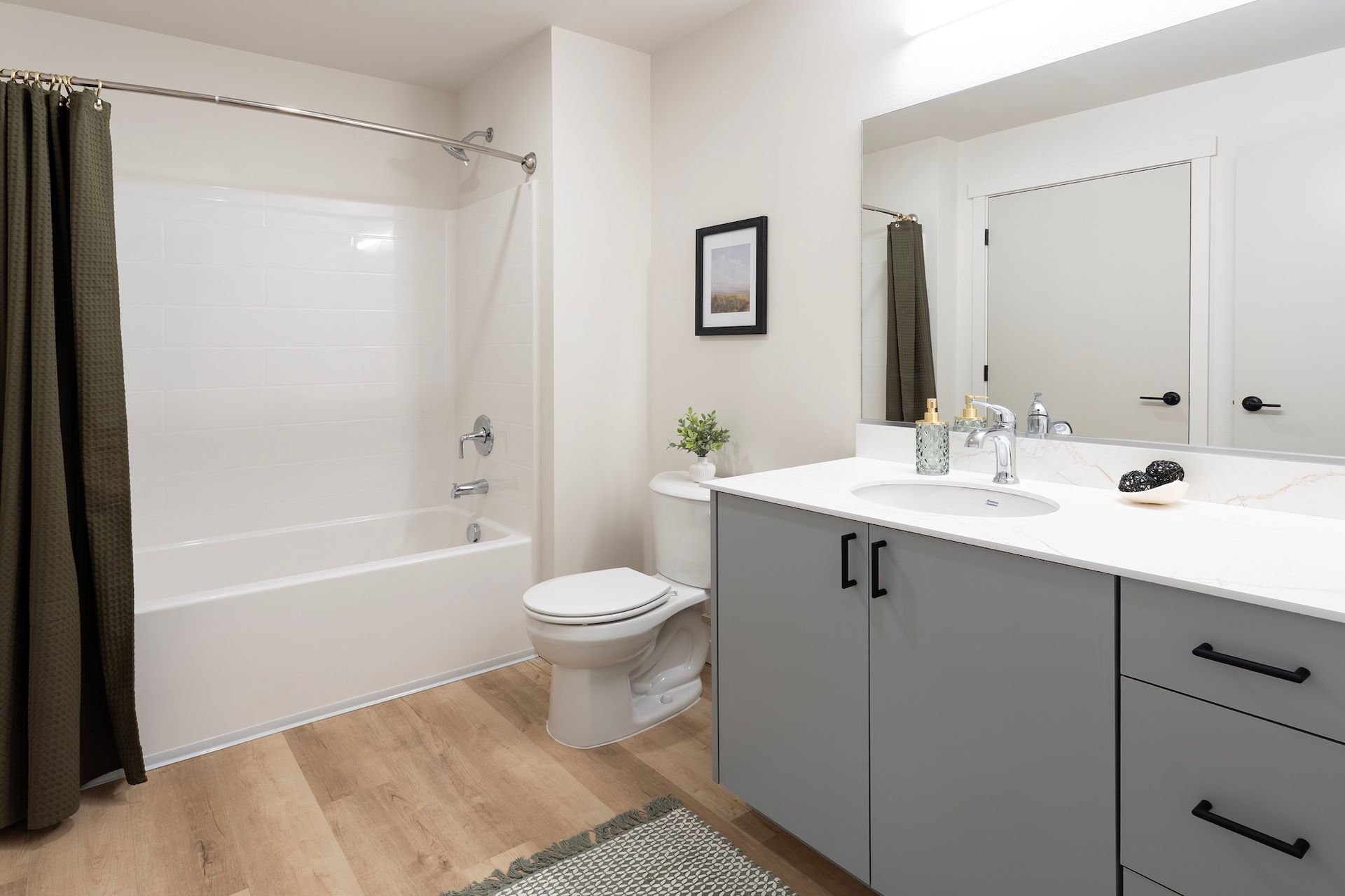 A modern bathroom featuring a white bathtub, toilet, and sink with gray cabinets. A green shower curtain and wooden floors are also visible.