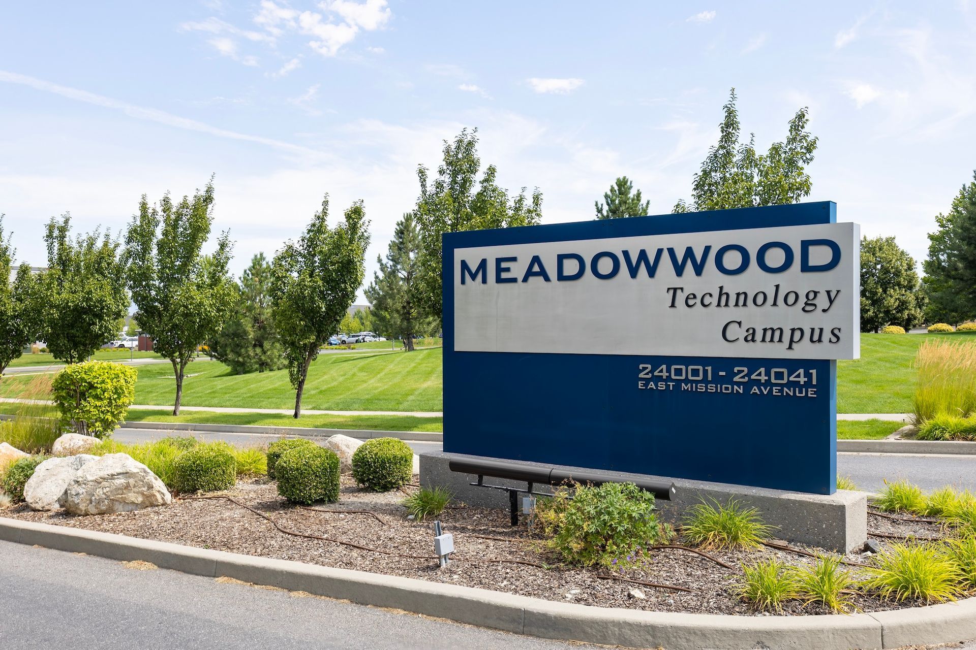 Meadowood Technology Campus sign in front of green bushes and trees under a bright blue sky.
