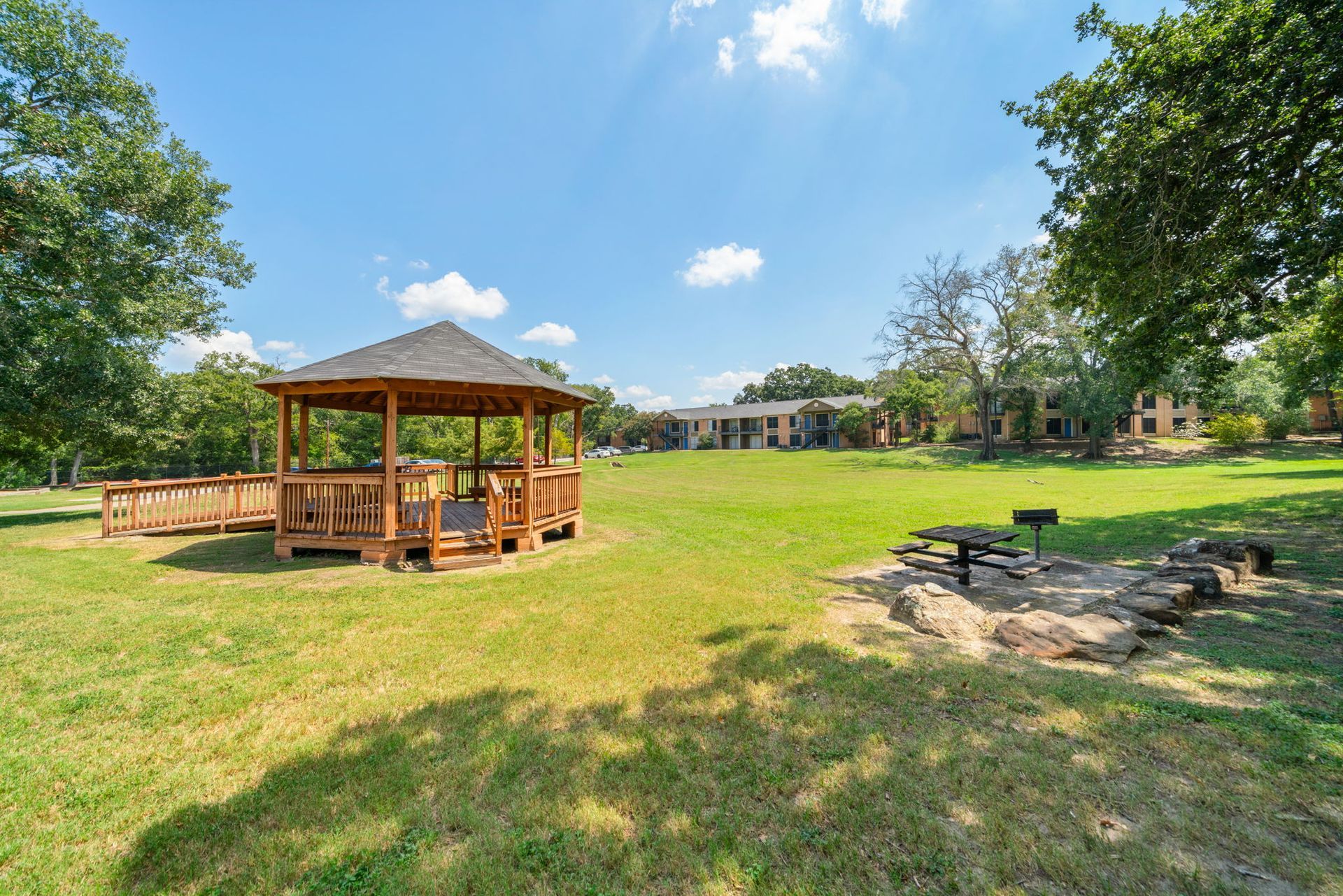 Wooden gazebo on green grass, with trees and apartment buildings in background under a sunny, blue sky.