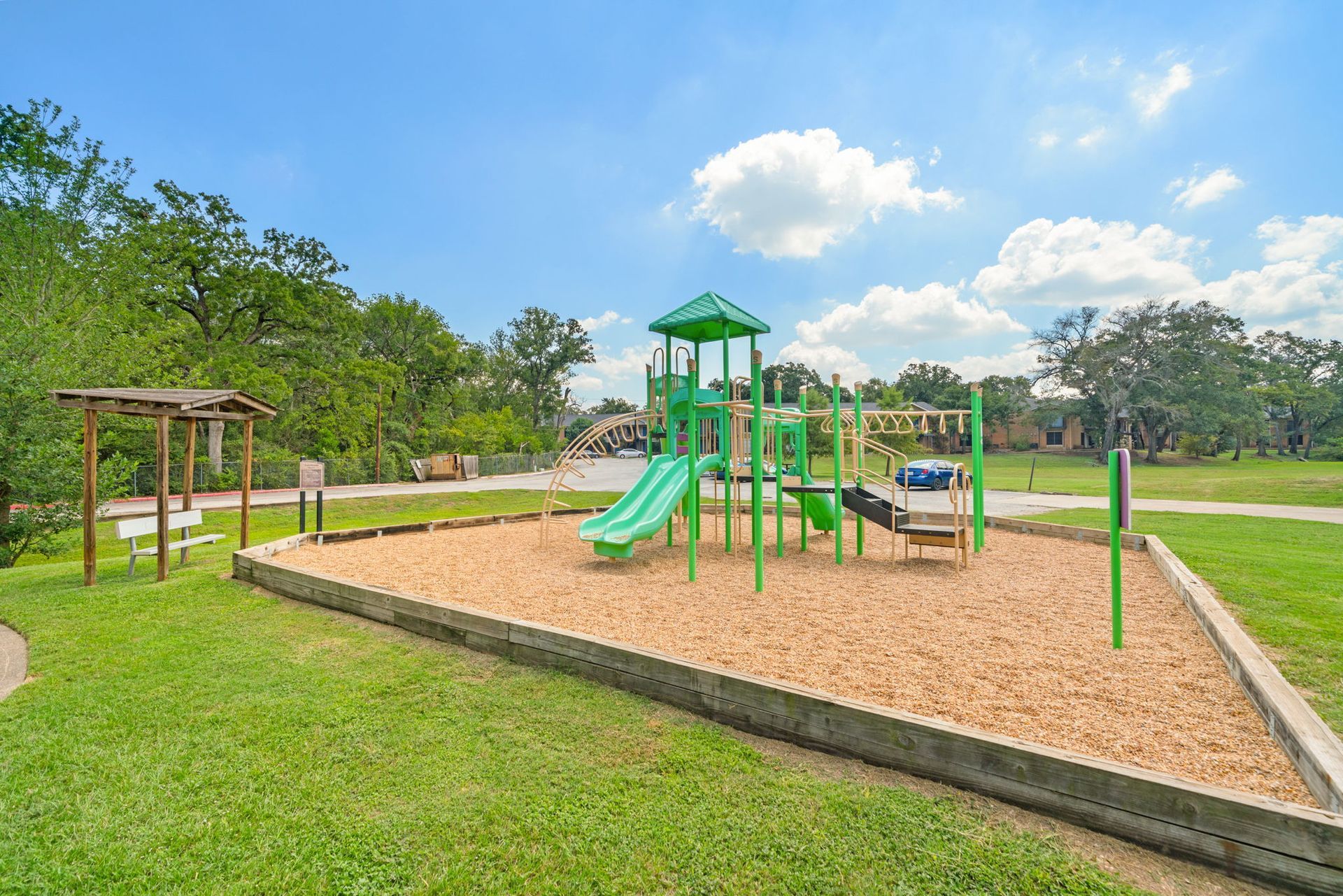 Playground with green equipment, slide, and wood chips, in a grassy park setting under a blue sky.
