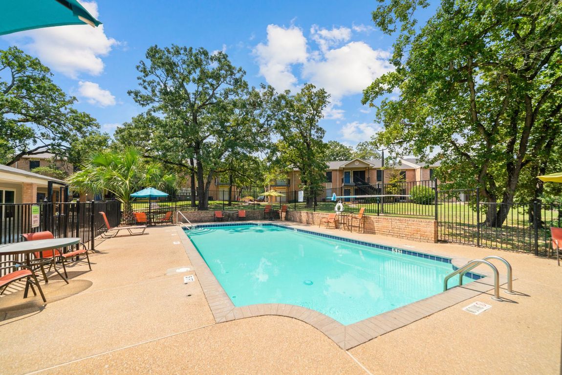 Swimming pool surrounded by lounge chairs, trees, and buildings on a sunny day.