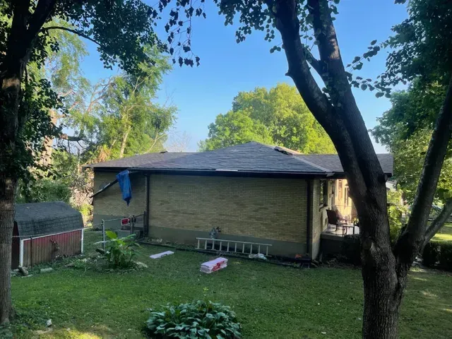 A brick house with a dark roof, surrounded by green trees and a grassy yard.