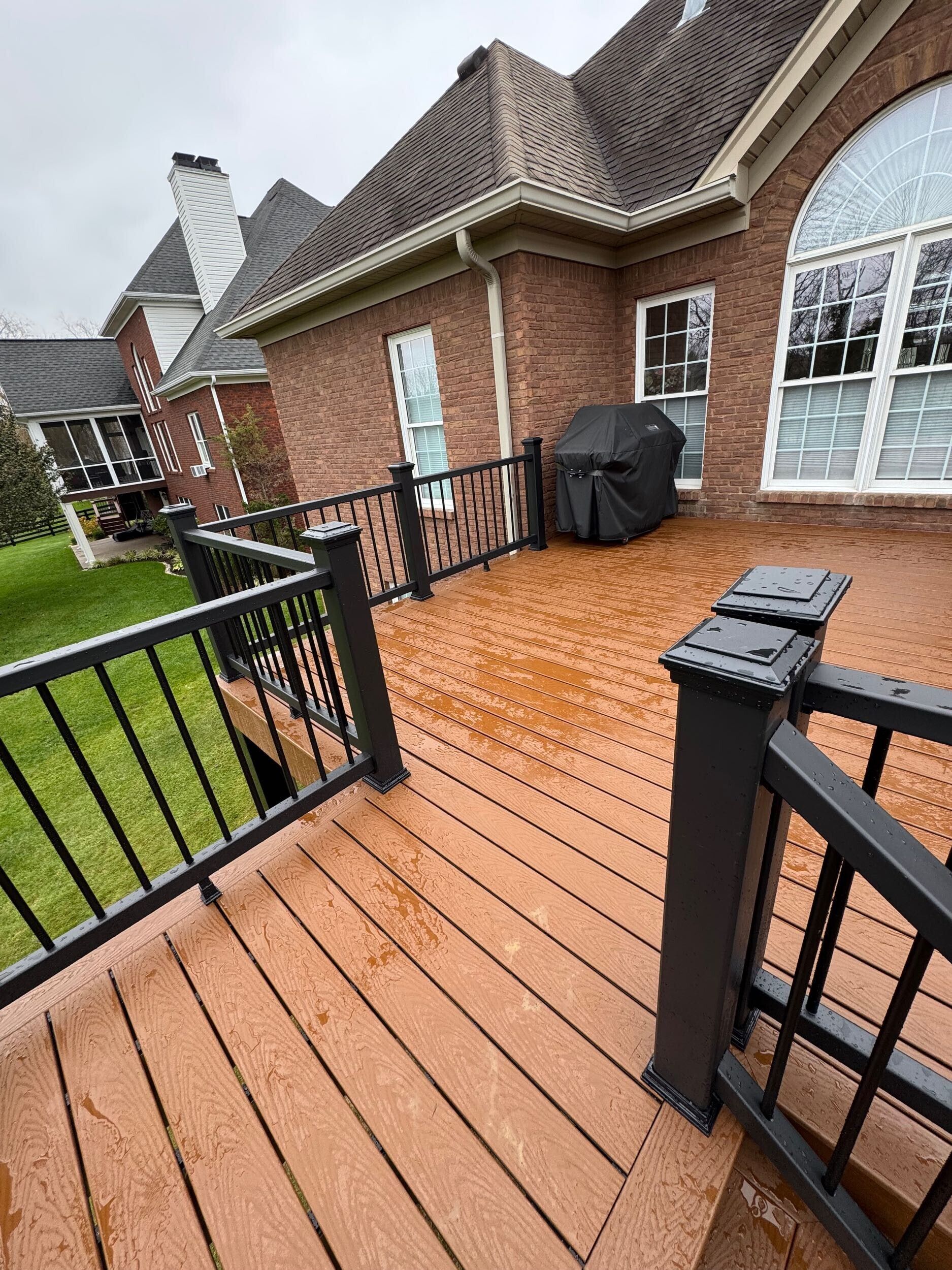 Wooden deck with black railings and brick house exterior; grill covered.