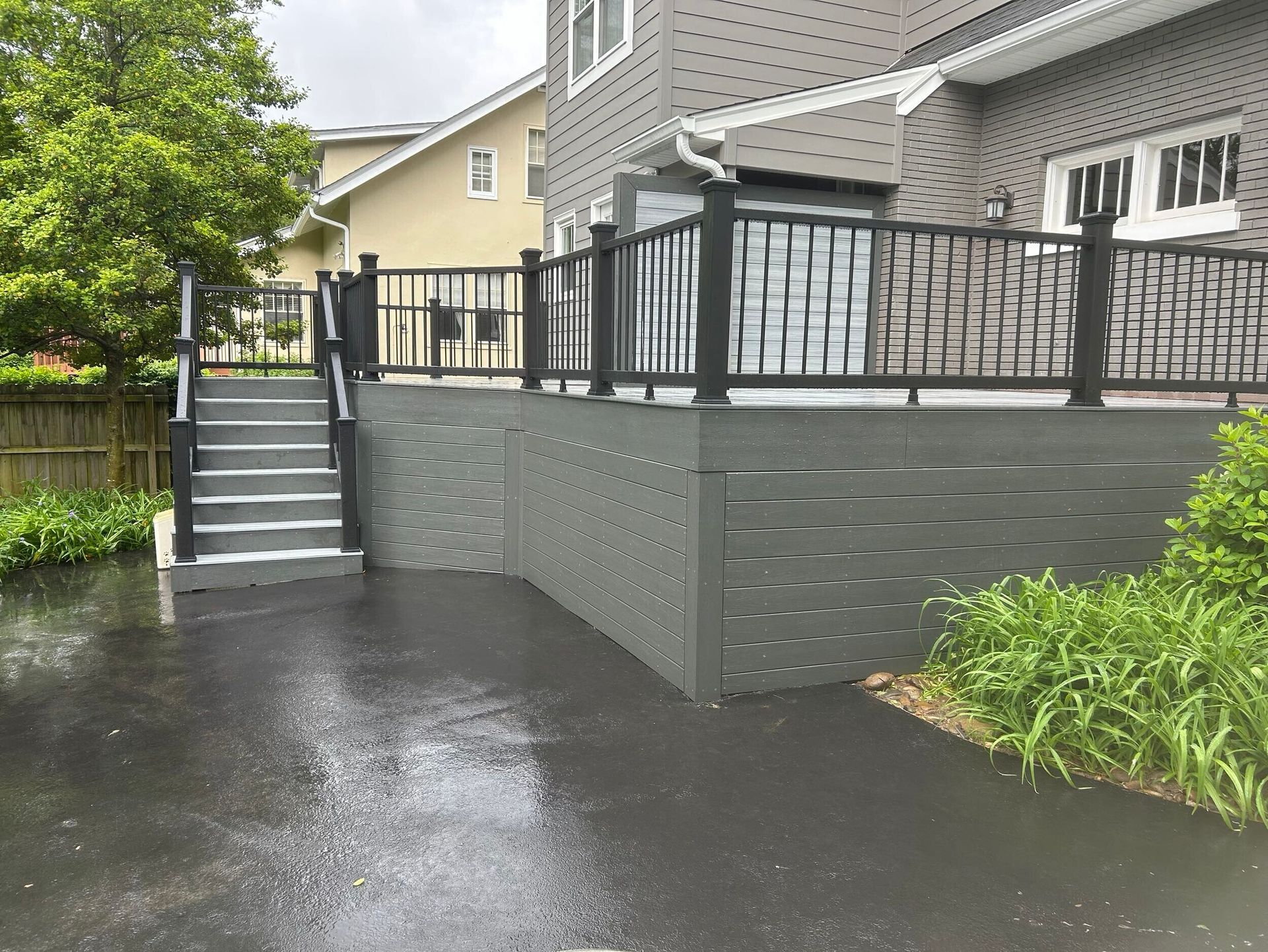 Grey composite deck with black railings and steps next to a house with dark siding. Wet asphalt driveway.