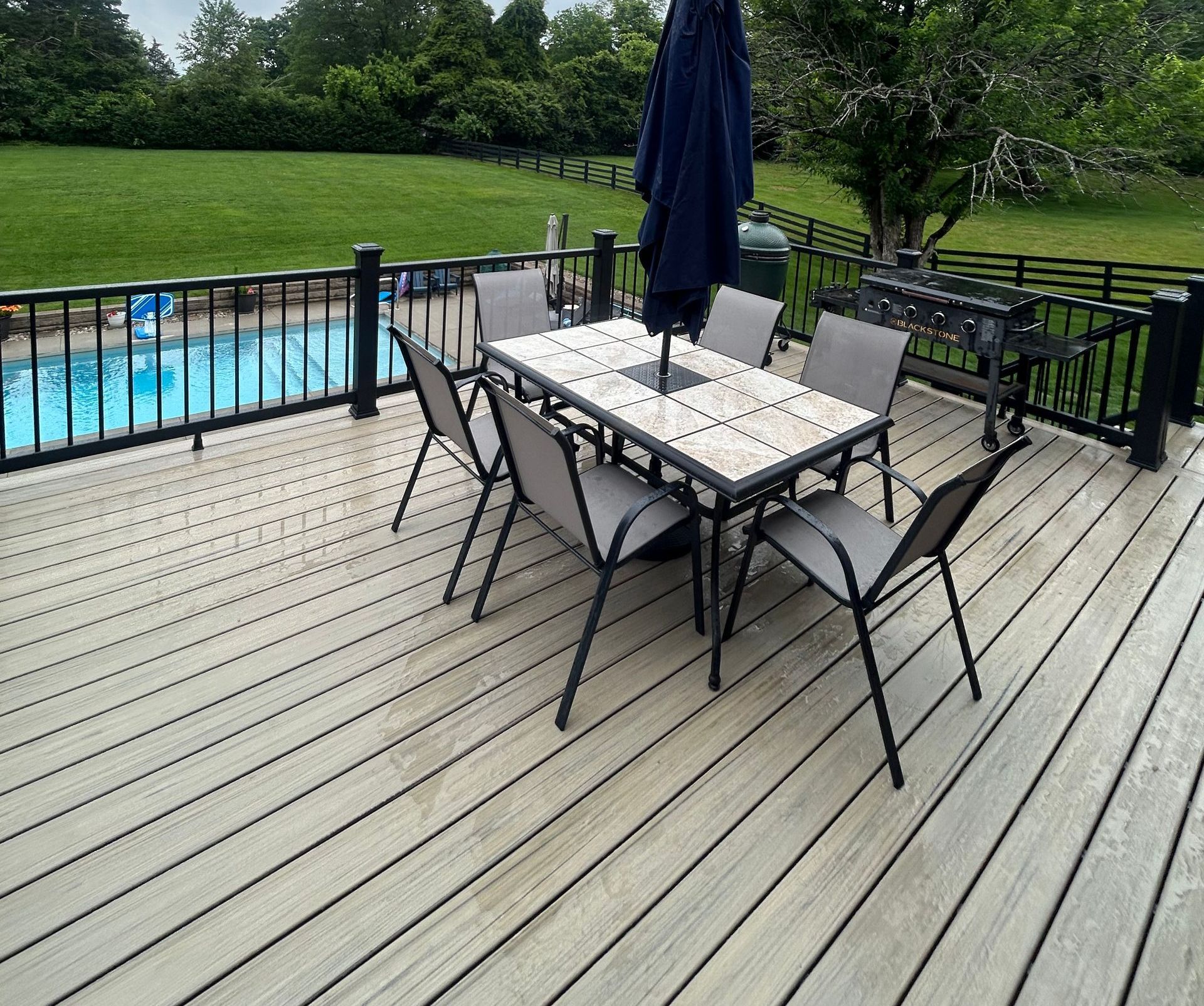 A deck with a table, chairs, and umbrella overlooks a pool and grassy yard with a black fence.
