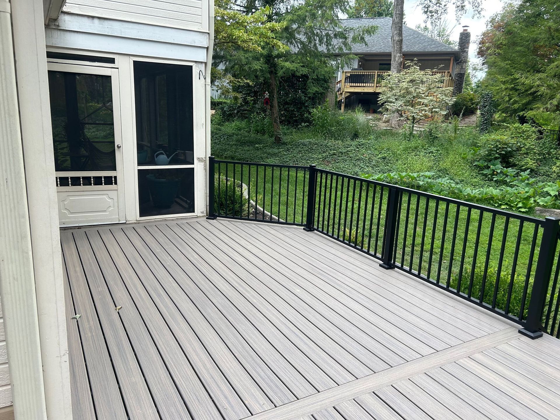 Gray composite deck with black railing next to a screened porch, overlooking a green, sloped yard.