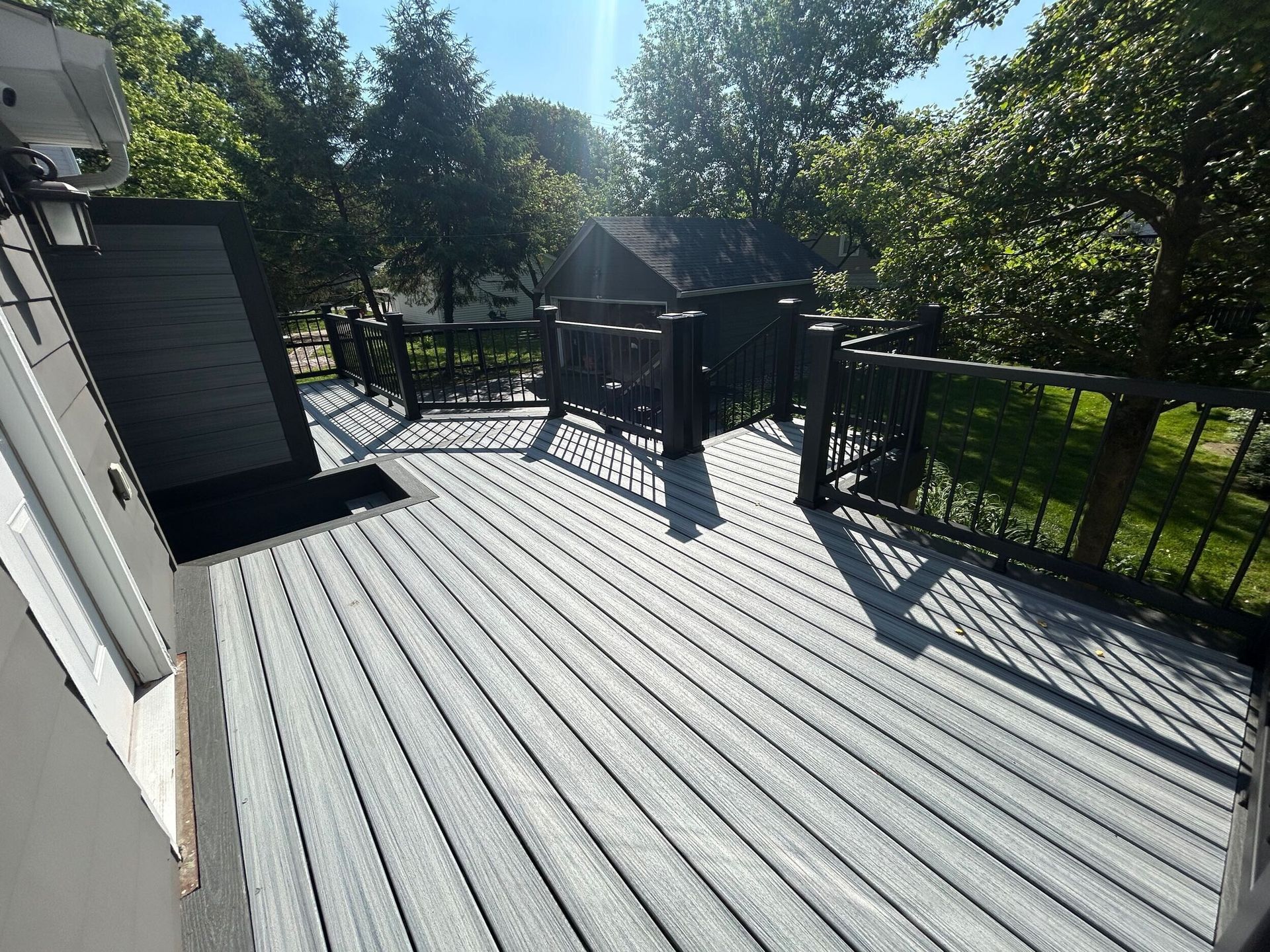 Grey deck with black railing surrounded by trees on a sunny day.