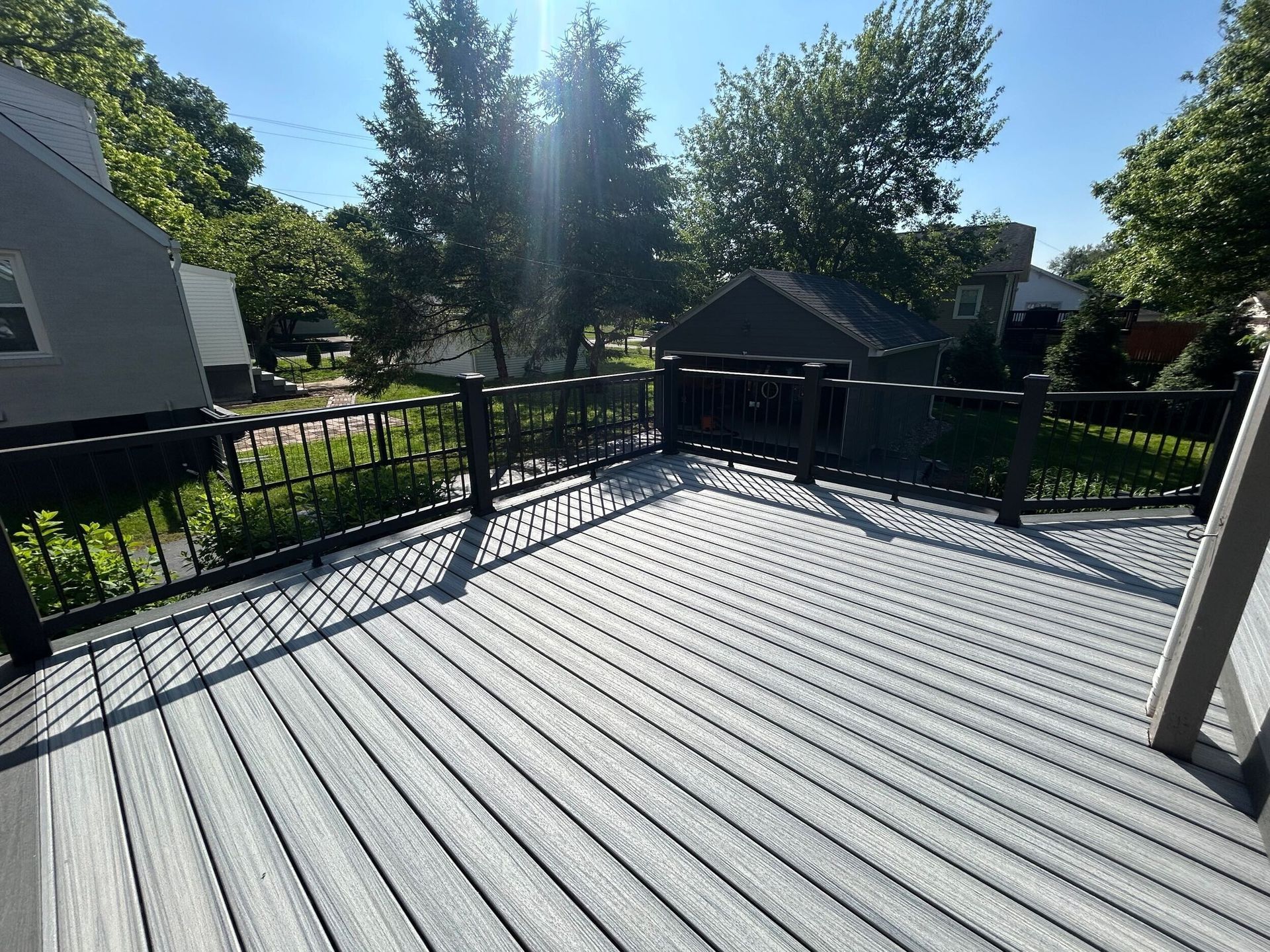 Gray composite deck with black railing on a sunny day. Backyard with trees and a small shed in the background.