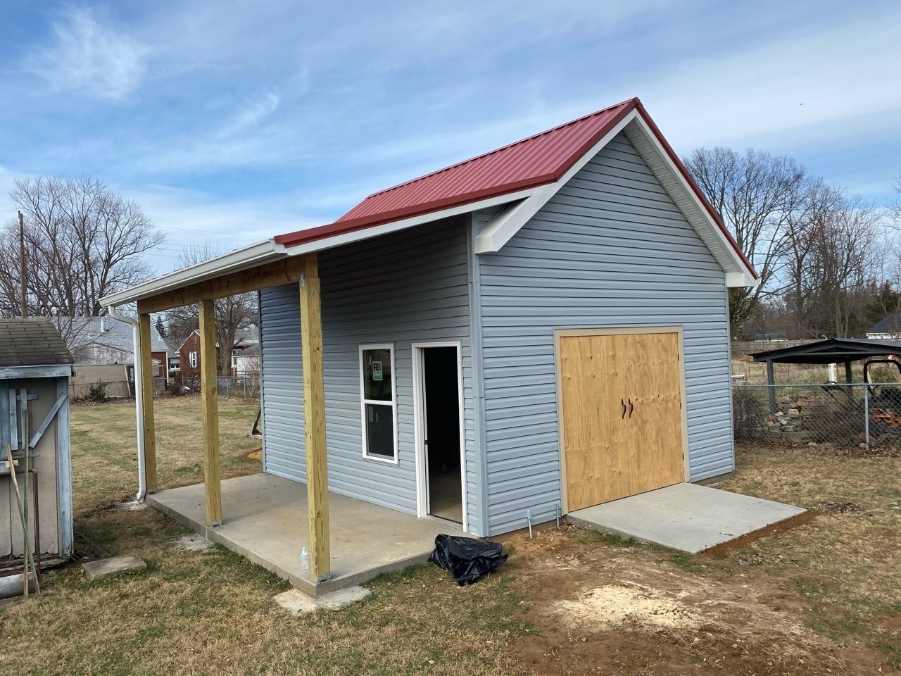 Small blue building with red roof, wooden door and porch. Set in a yard, cloudy sky.