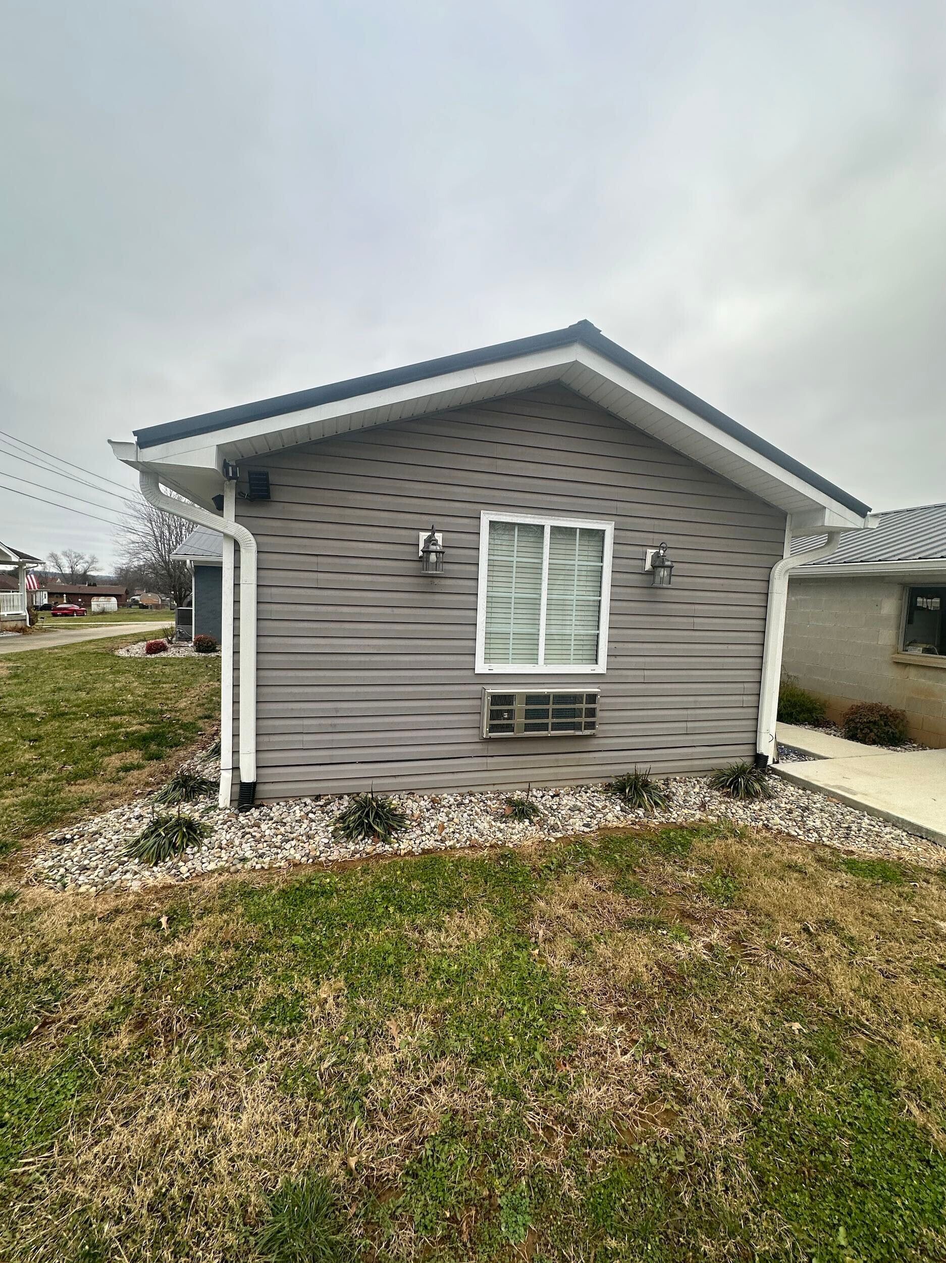 Small gray building with a white-trimmed window, two sconces, and a dark roof. Gray siding and a stone bed.