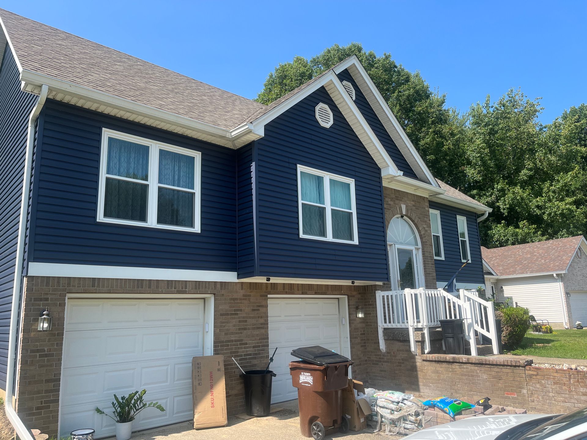Two-story house with dark blue siding and brick facade. White trim, two-car garage, and blue sky.
