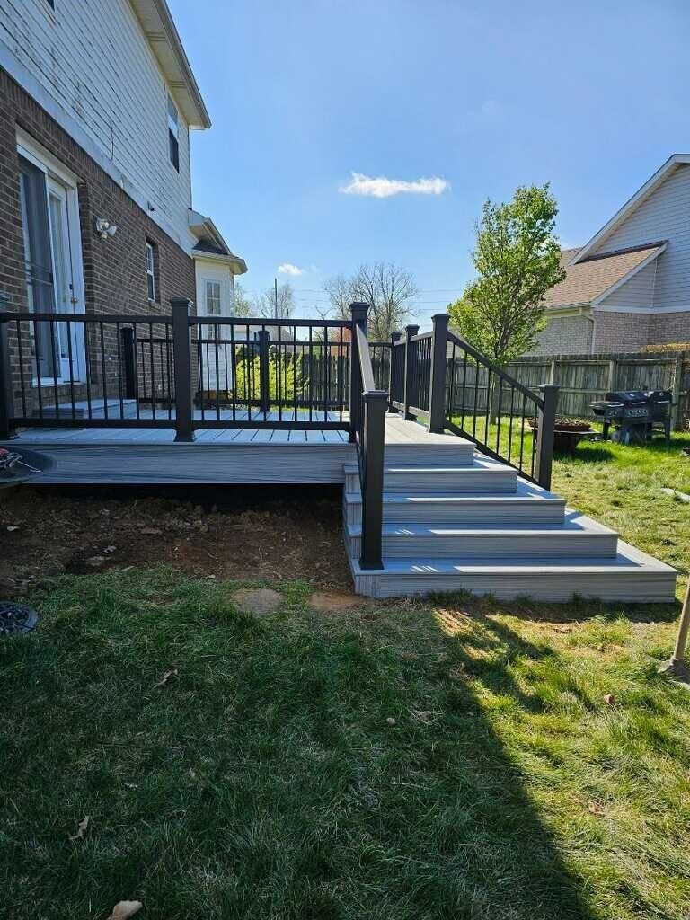 Gray deck with black railing and steps leading to a backyard. Sunny day.