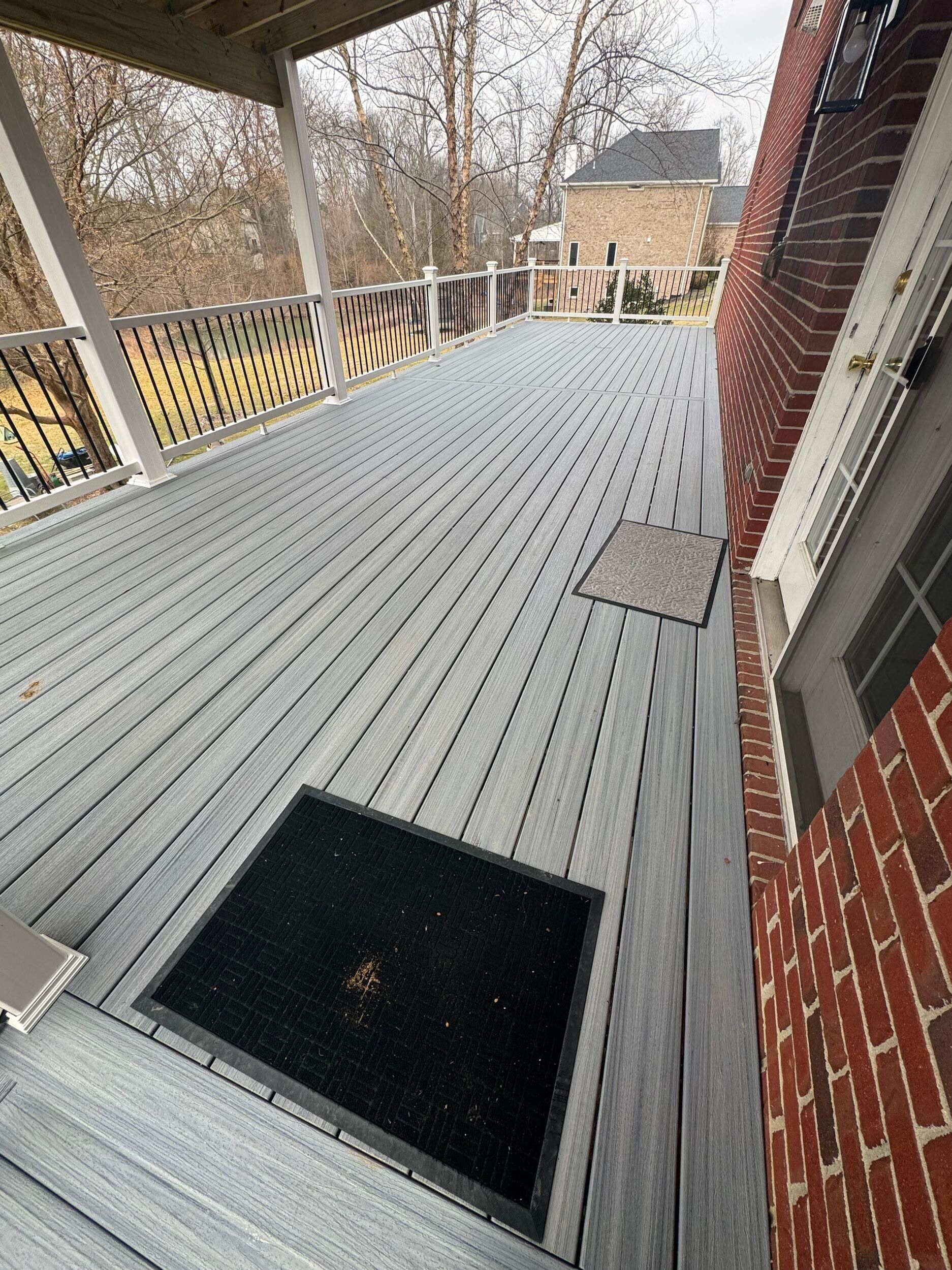 A gray composite deck with a black mat is next to a red brick house; a wooden railing is on the left.