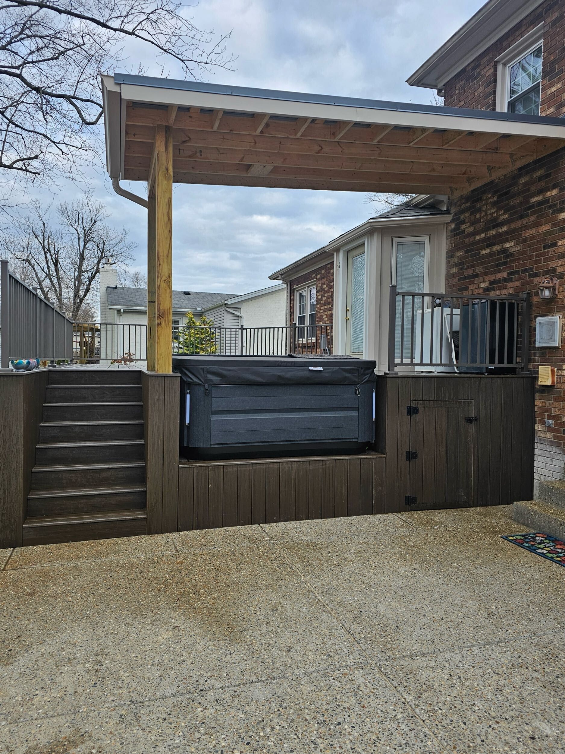 Hot tub on a wooden deck with a covered patio, next to a brick building and gravel.