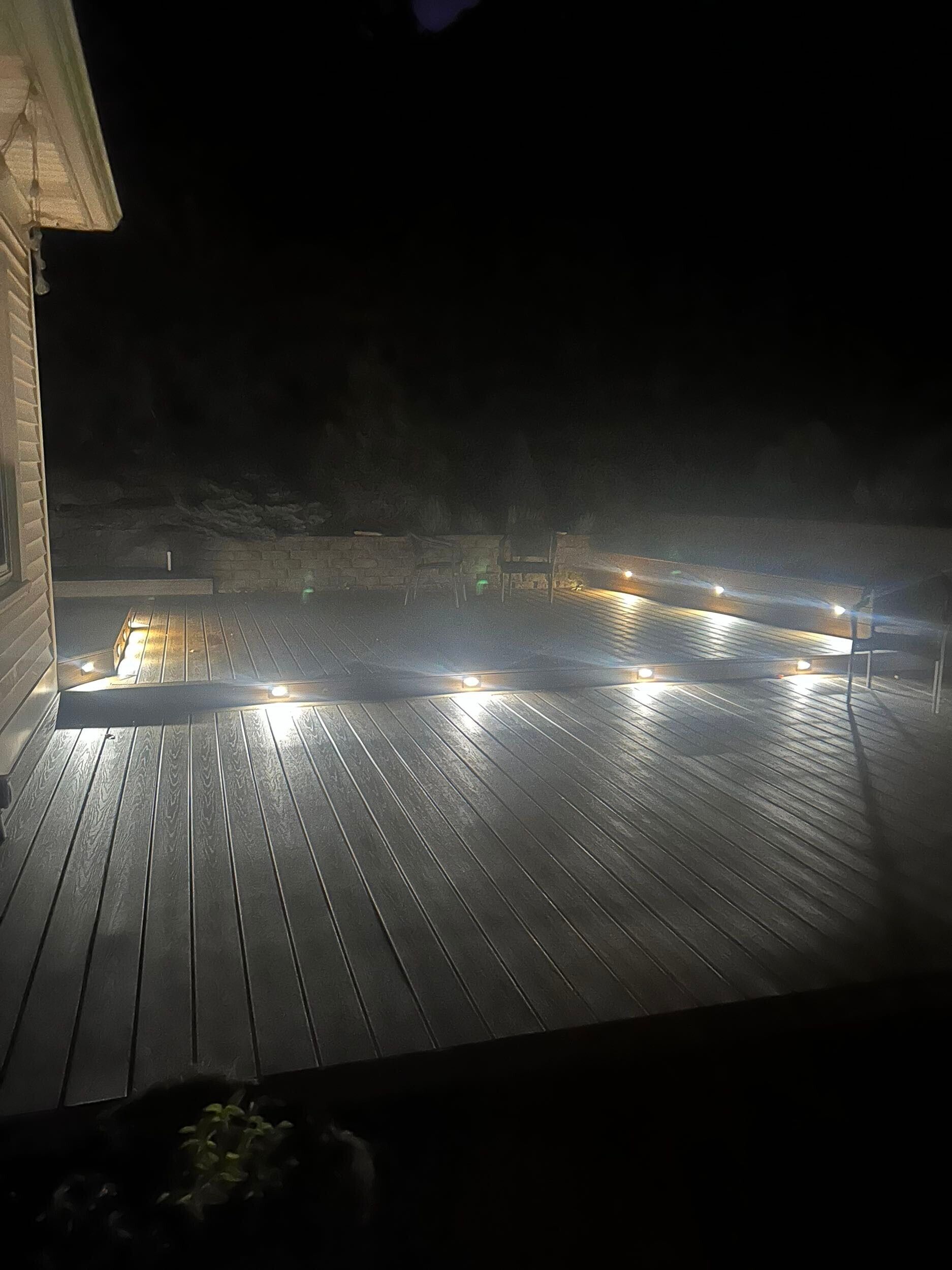 Nighttime view of a wooden deck lit by several lights, attached to a house with dark background.