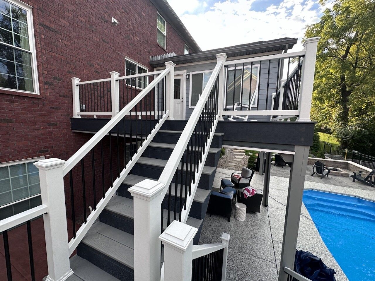 Deck with stairs descending to a poolside patio; white and black railing, brick exterior, trees visible.
