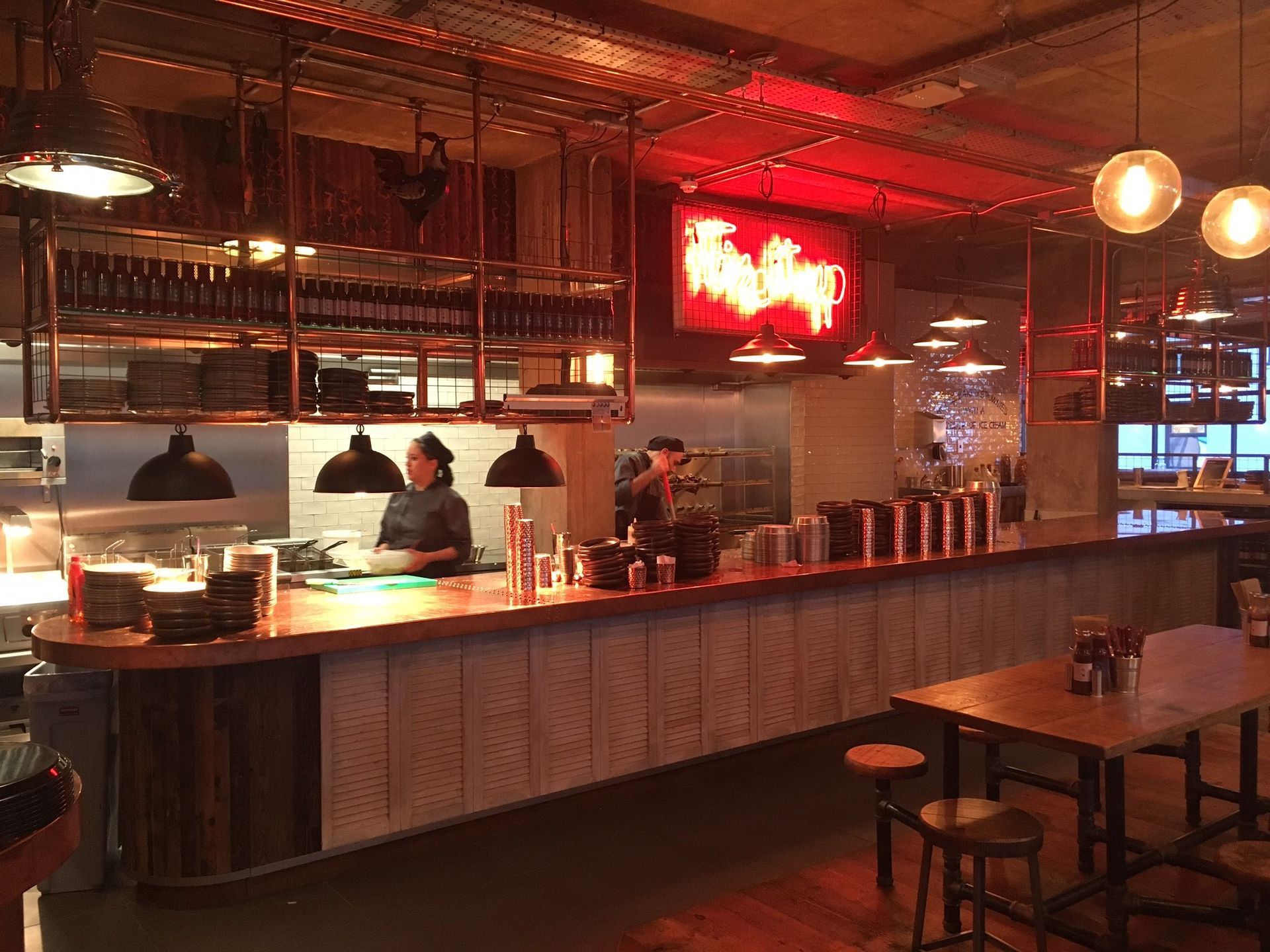 Chicken Society, Finchley, London, looking towards kitchen pass counter with louvre panels to counterfront, industrial pendant lights, crockery gantry, and feature neon sign.