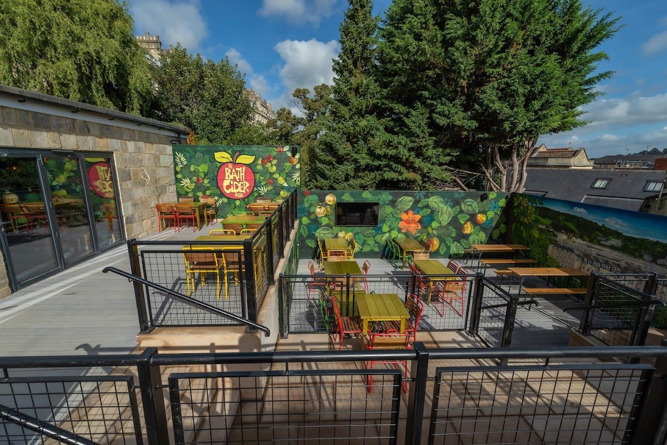Bath Cider House, view of rear terraced garden area, with decking, coloured furniture, and feature garden mural to perimeter scaffold board fencing.