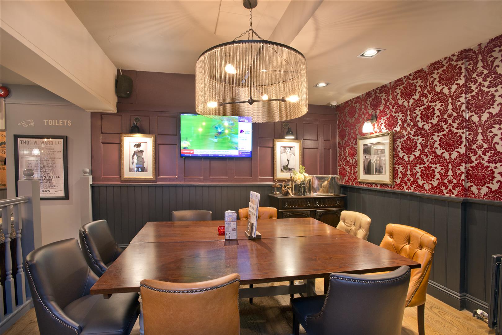 The Old Brewery, Marlow, opulent rear raised dining area with large dining table and large feature light overhead, timber panelled walls and decorative wallpaper.