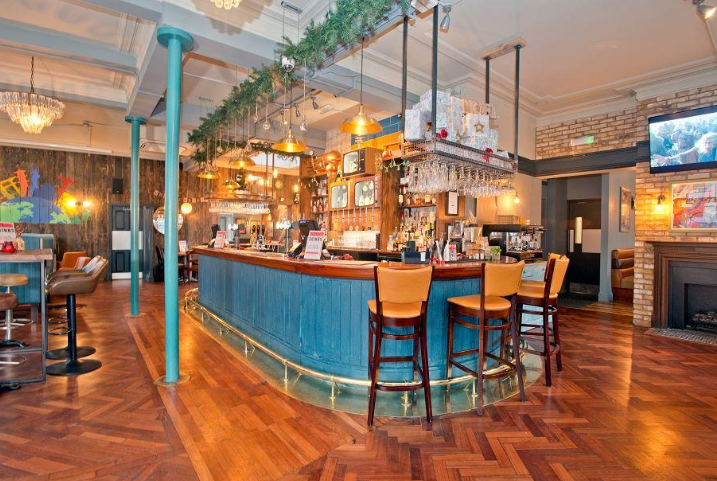 Whitecross Tap, Old Street, London, ground floor, looking towards main bar counter, timber panelled painted bar front and overhead pendant lights, with retro TV feature bottle displays to backfitting.