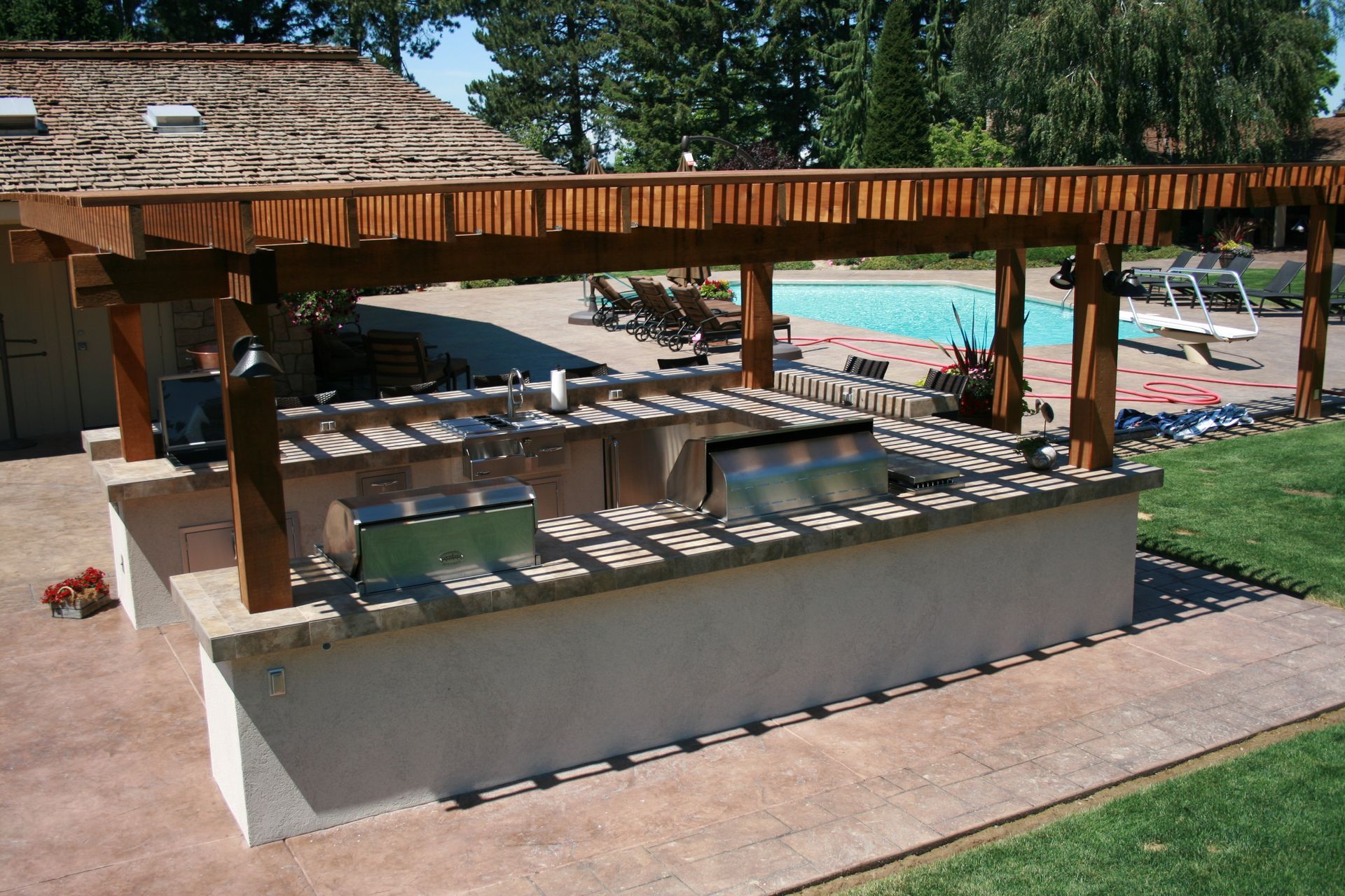 A large outdoor kitchen under a pergola with a pool in the background