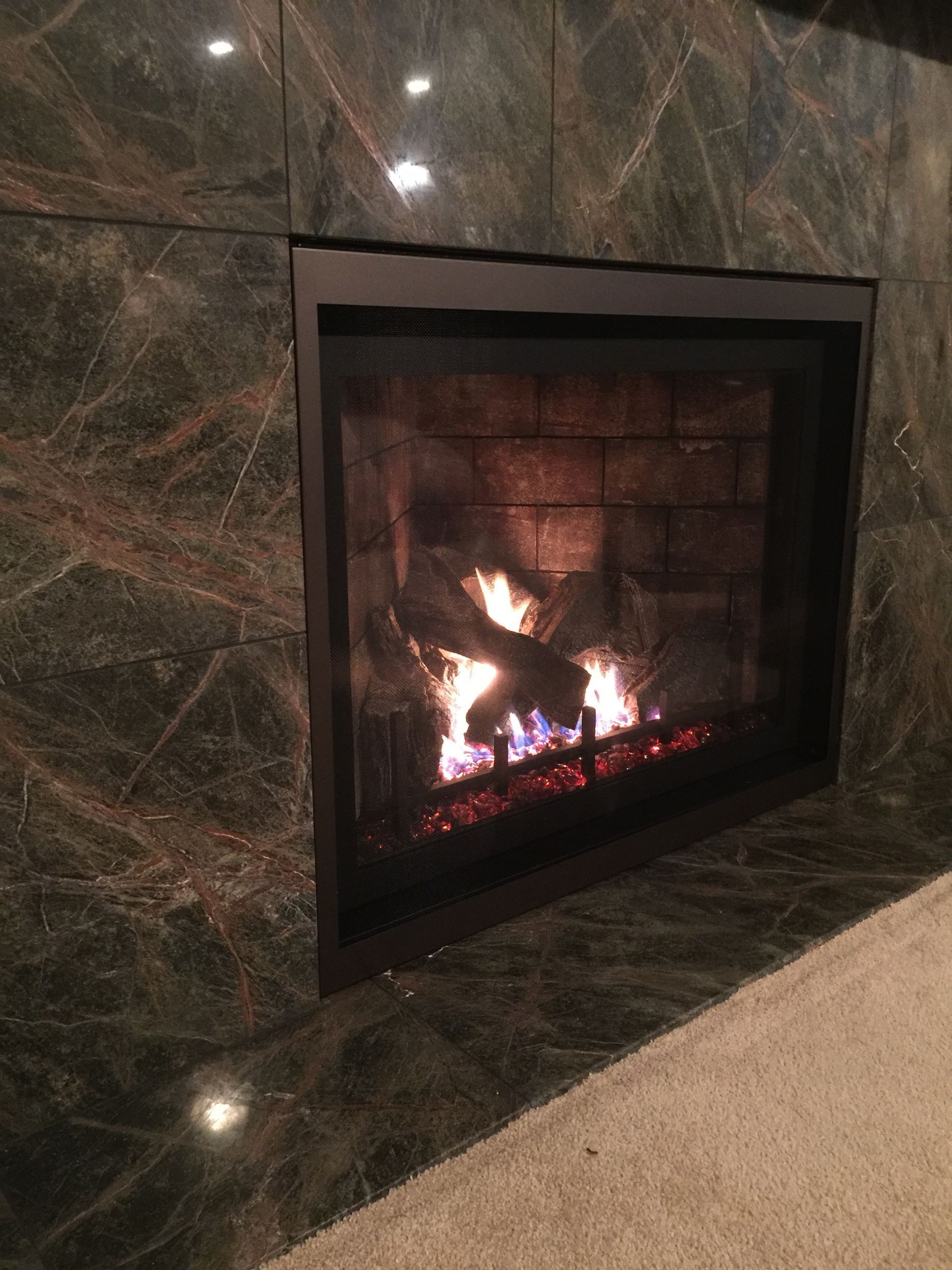 A fireplace is lit up in a room with a marble wall.