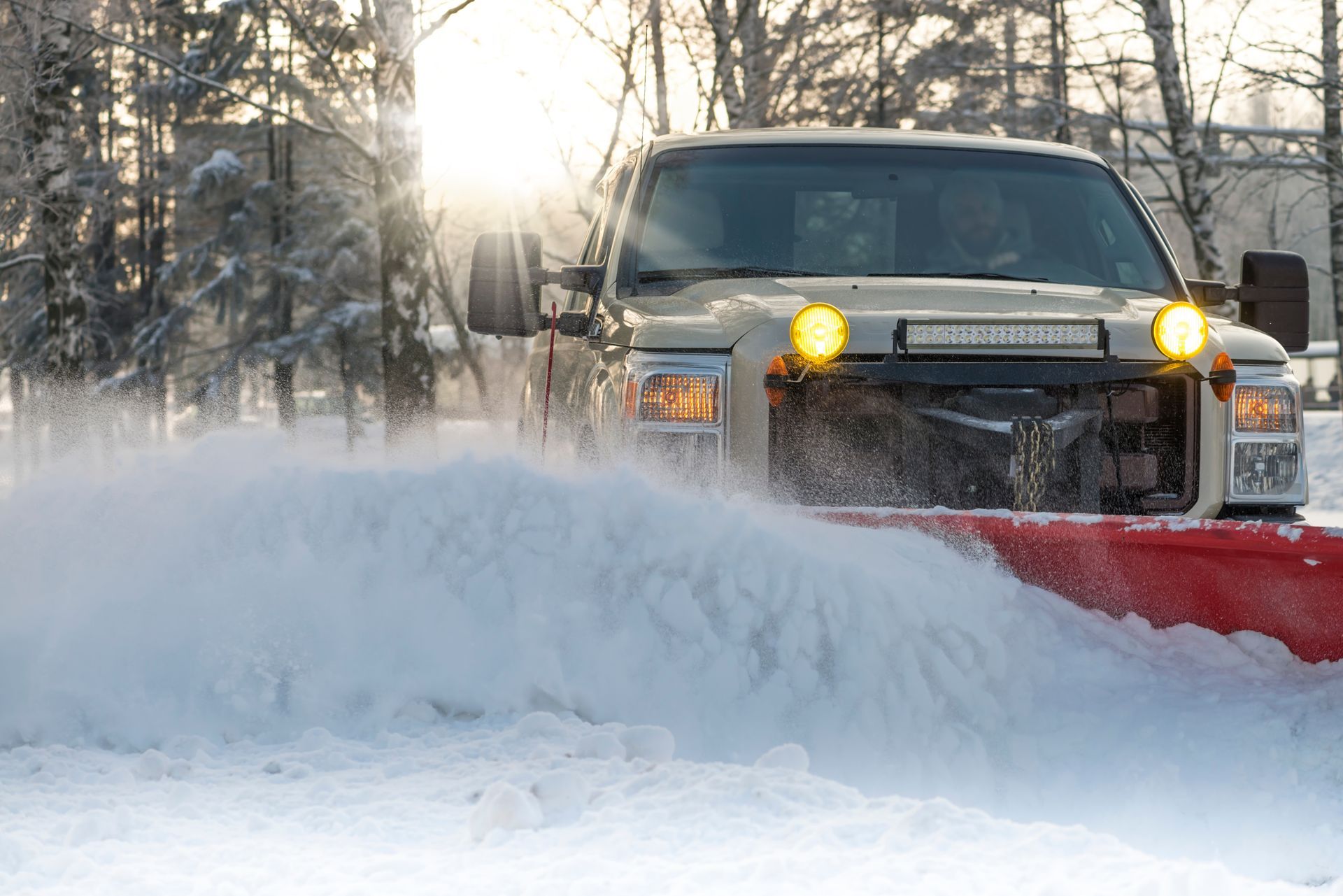Truck with plow clearing snow, headlights on in a snowy forest setting.