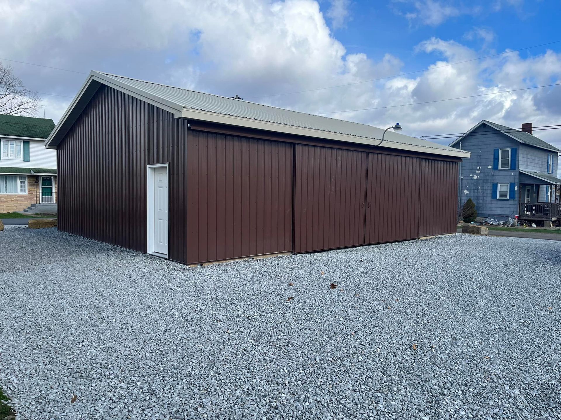 A brown garage with a white door is sitting in a gravel lot next to a house.