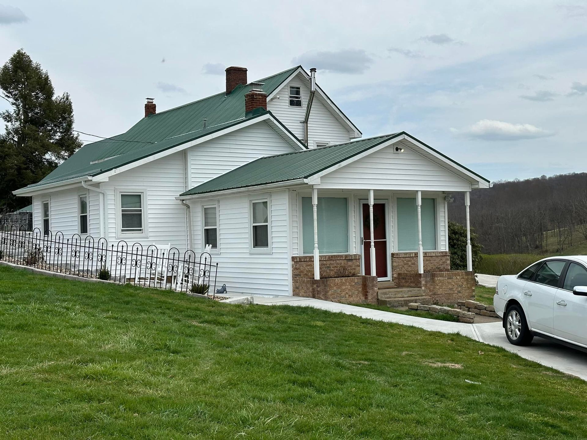 A white car is parked in front of a white house with a green roof.